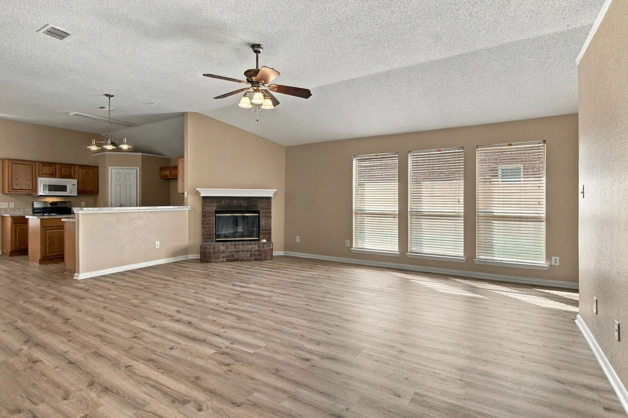 Wood-finish flooring throughout, brick fireplace with white mantel, kitchen featuring wood cabinetry and white appliances, multiple windows, and a ceiling fan with integrated lighting