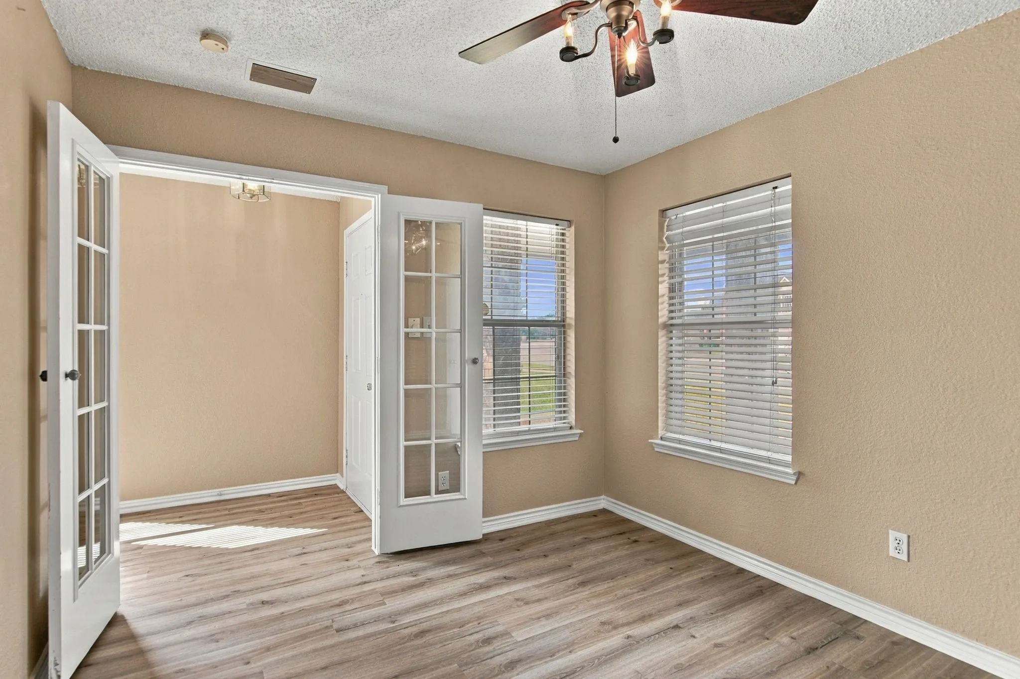 Wood-finish flooring, two windows with blinds, ceiling fan with integrated lighting, and double French doors with clear glass panels