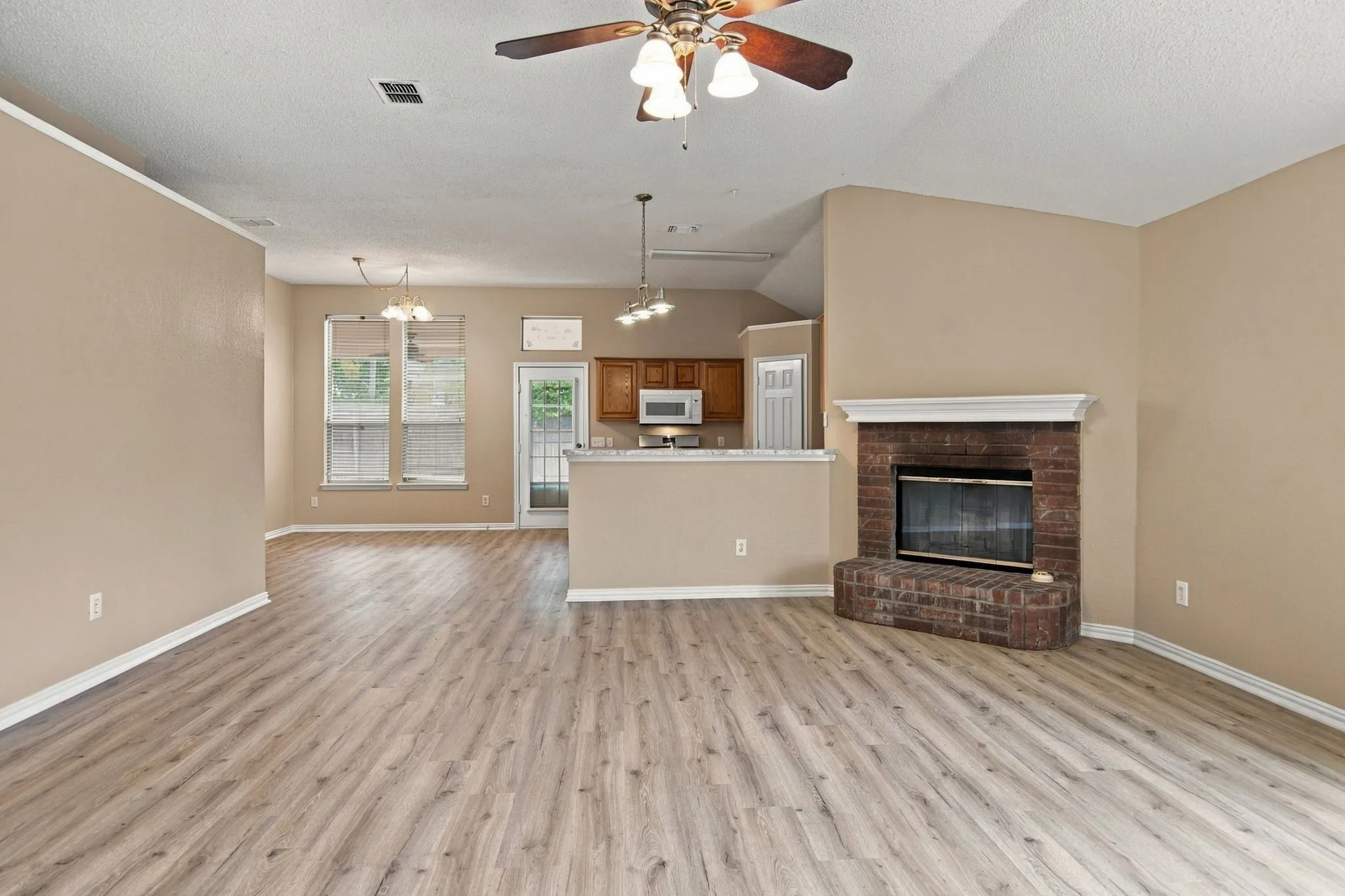 Vaulted ceiling with a ceiling fan, wood-finish flooring, brick fireplace with a white mantel, kitchen with wood cabinetry, and a double window