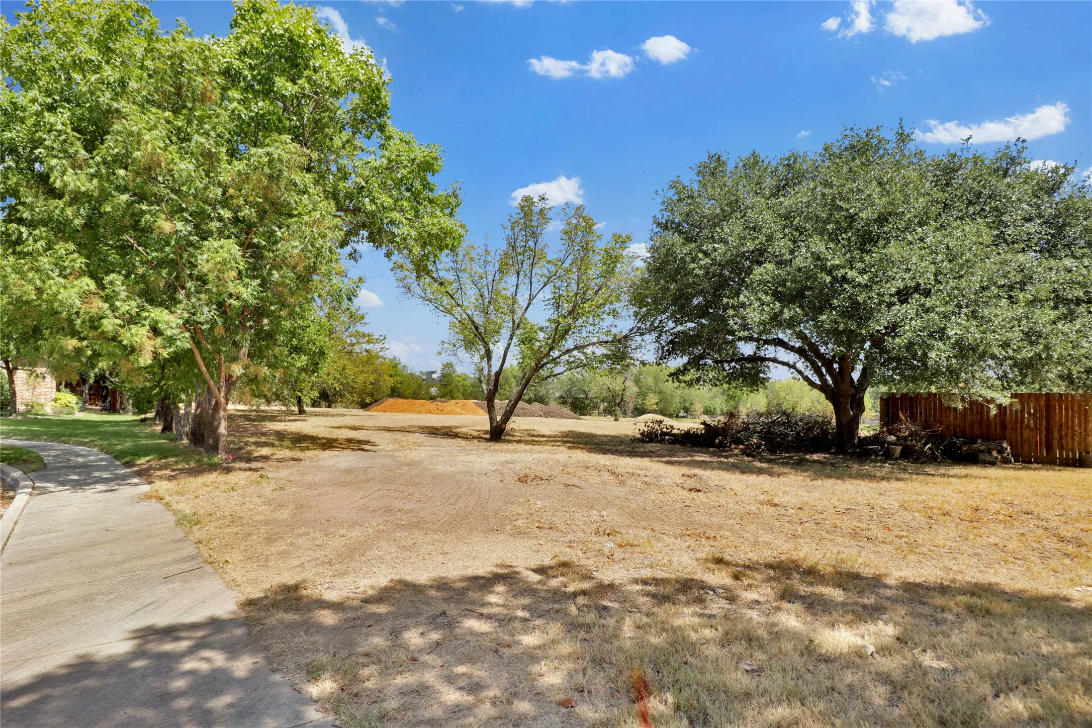Expansive land parcel featuring mature trees, a paved walkway, and a partial wood fence