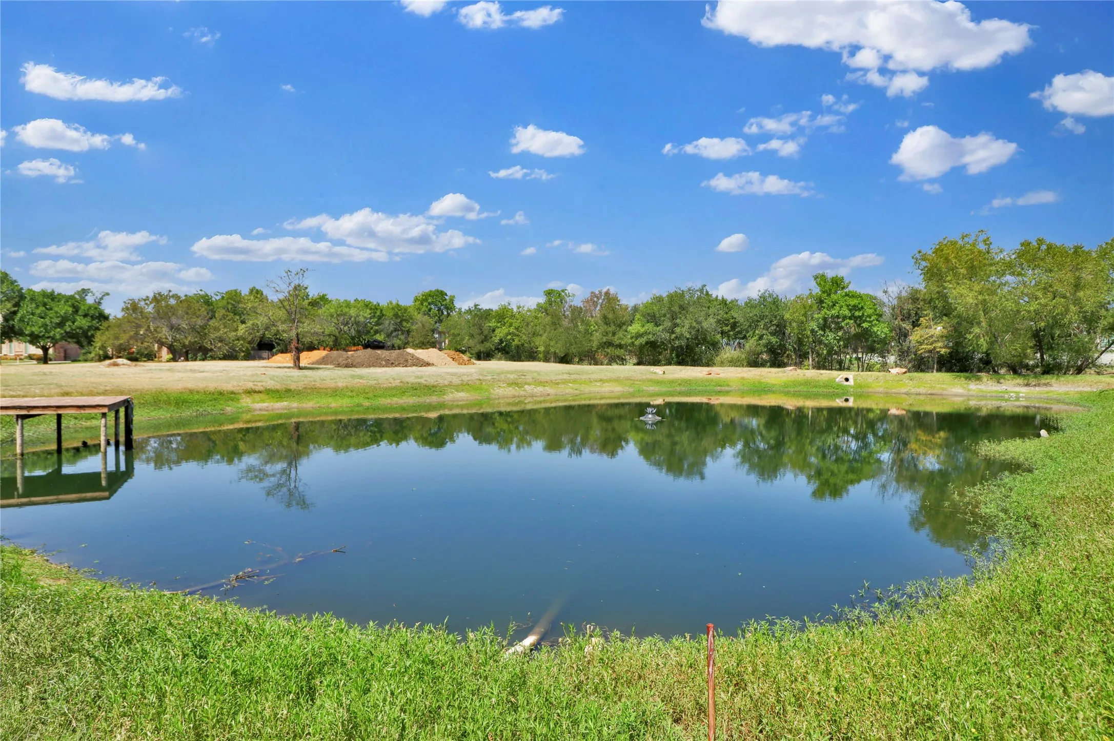 Expansive pond with tranquil water, surrounded by lush green landscaping and mature trees, featuring a wooden dock