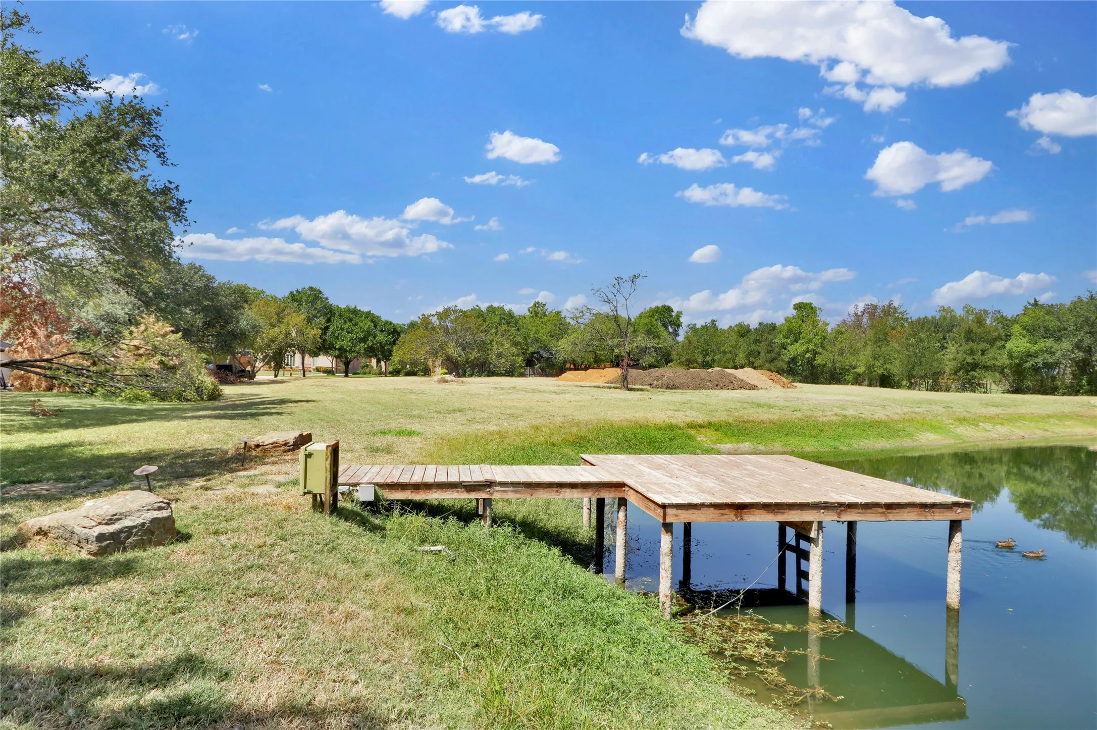Expansive outdoor area featuring a pond with a wooden dock, large grass areas, and mature trees
