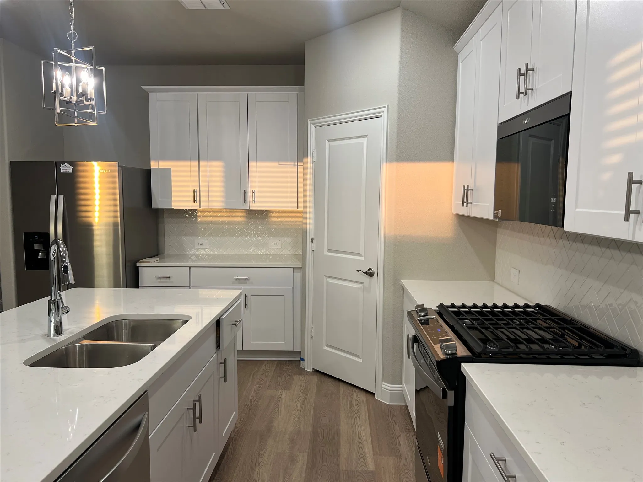 Kitchen featuring white shaker cabinetry, stainless steel appliances, a center island with a double basin sink, light countertops, and wood-finish flooring