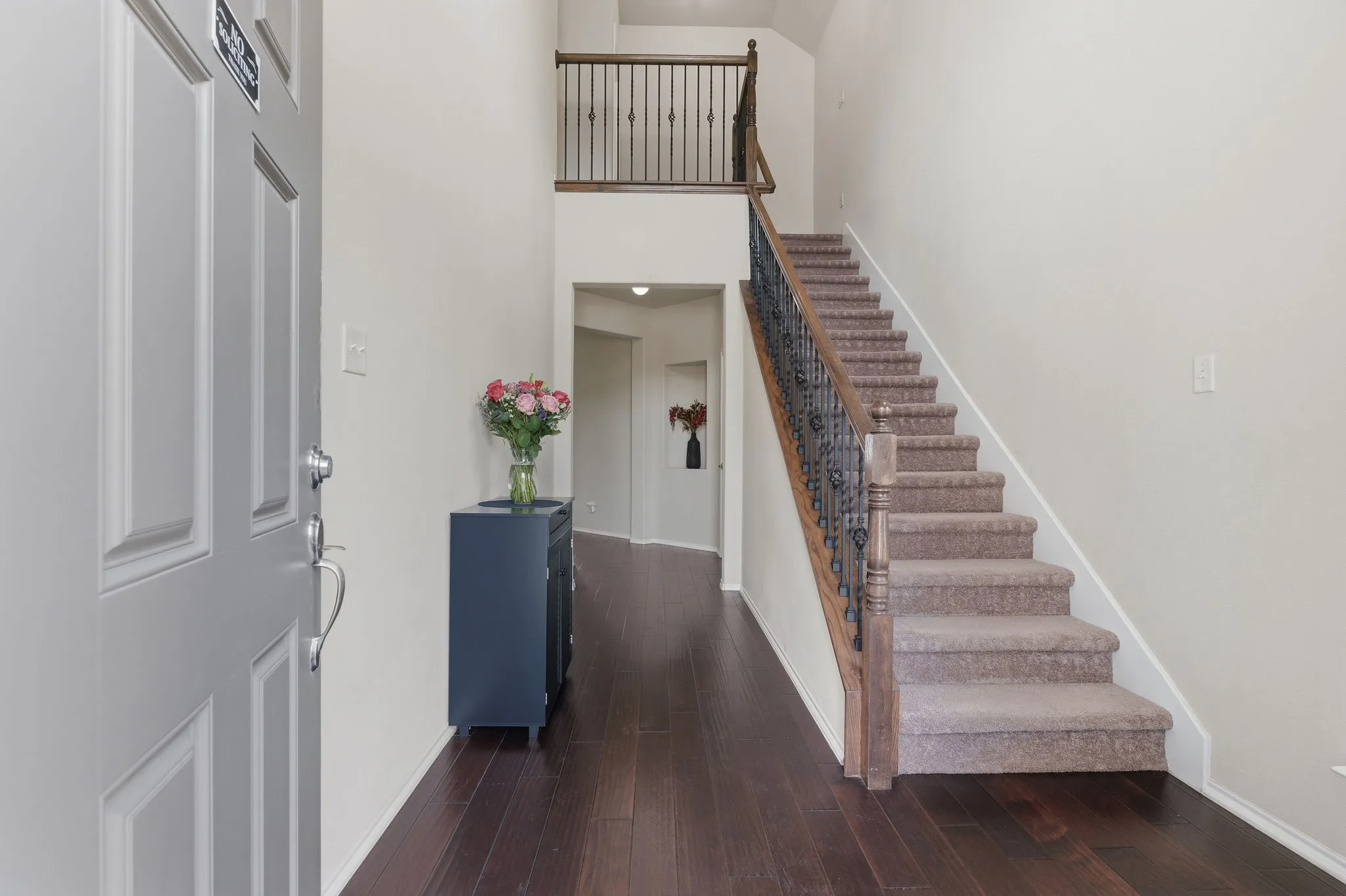 Dark wood-finish flooring extending into the hallway, a carpeted staircase with wood handrails, and decorative metal balusters