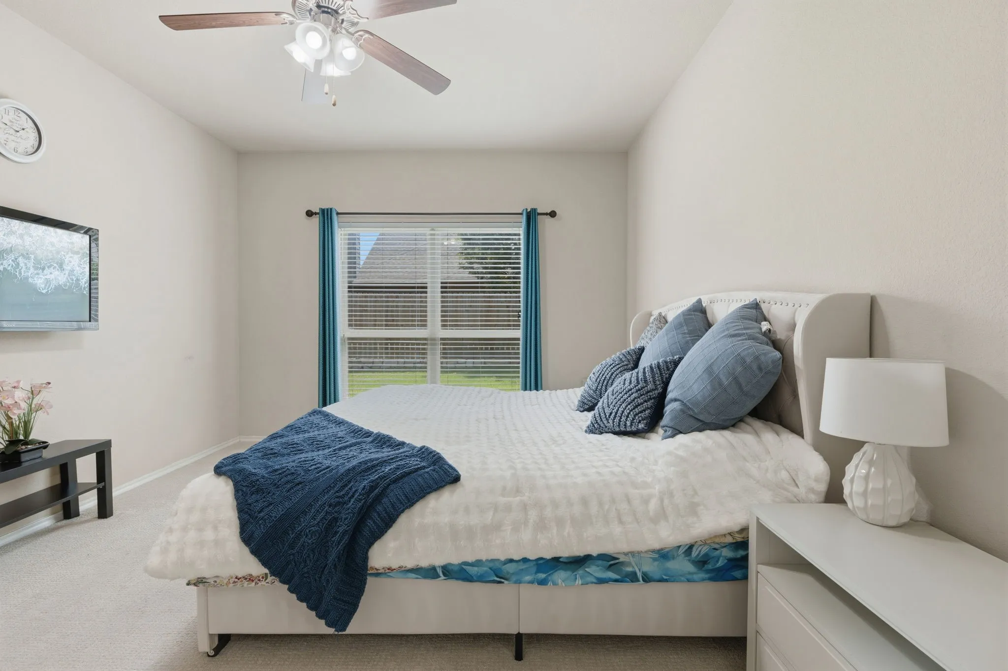 Ceiling fan with light fixture, window with blinds and curtains, wall-mounted television, upholstered headboard, and light-toned carpeting