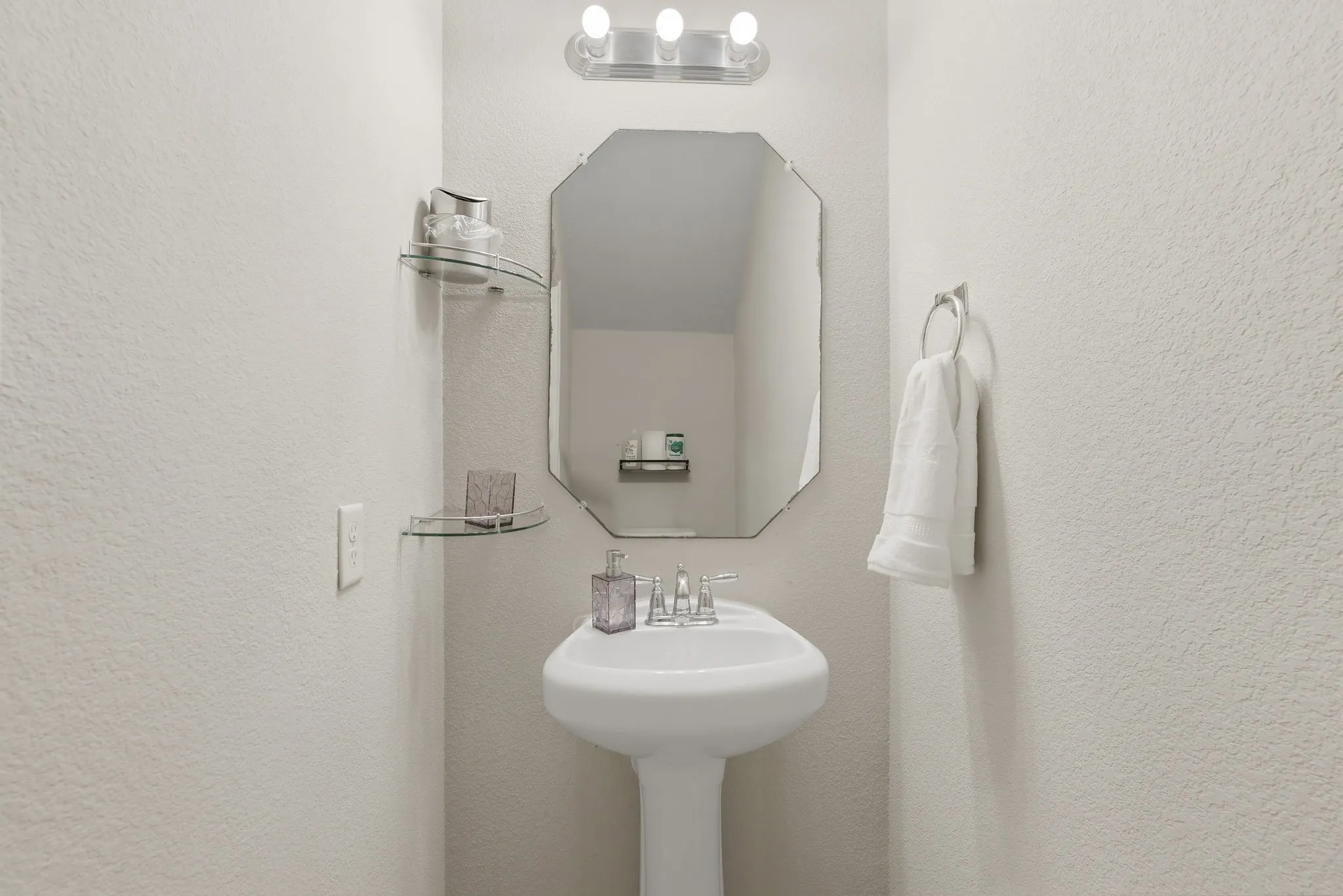 Pedestal sink with chrome faucet, an octagonal mirror, glass shelving, and a three-light vanity fixture