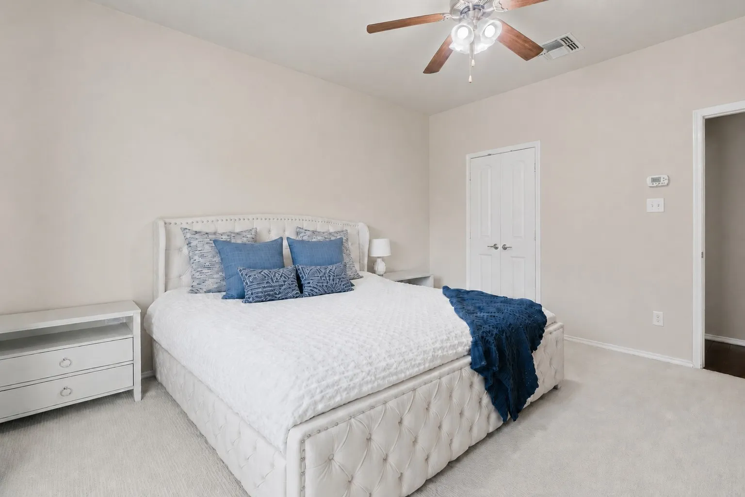 Neutral tone wall paint, carpeted flooring, white paneled doors, ceiling fan with integrated lighting, and a digital thermostat
