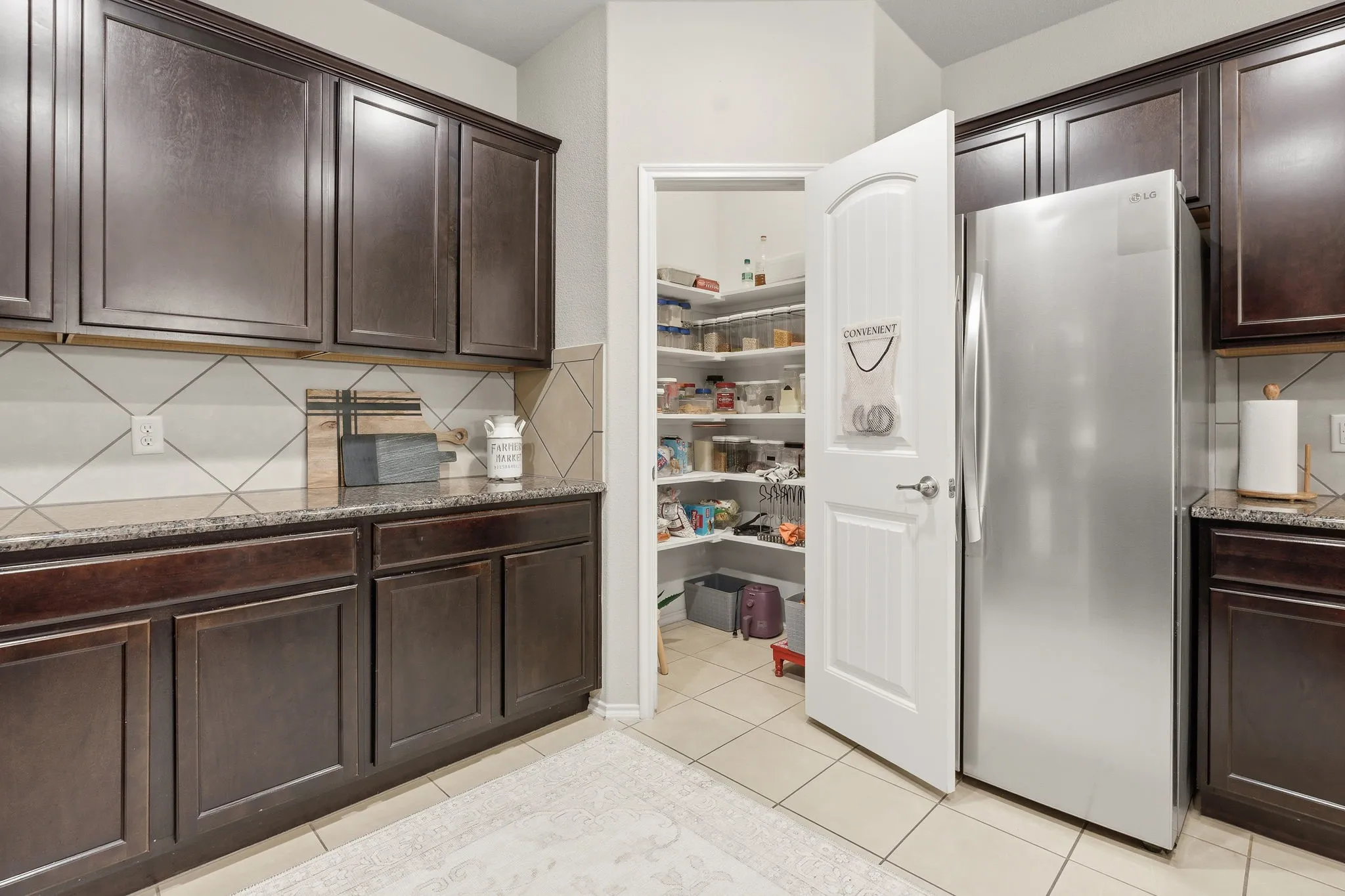 Dark wood-finish cabinetry, light-toned countertop, diagonal pattern tile backsplash, and a walk-in pantry with built-in shelving