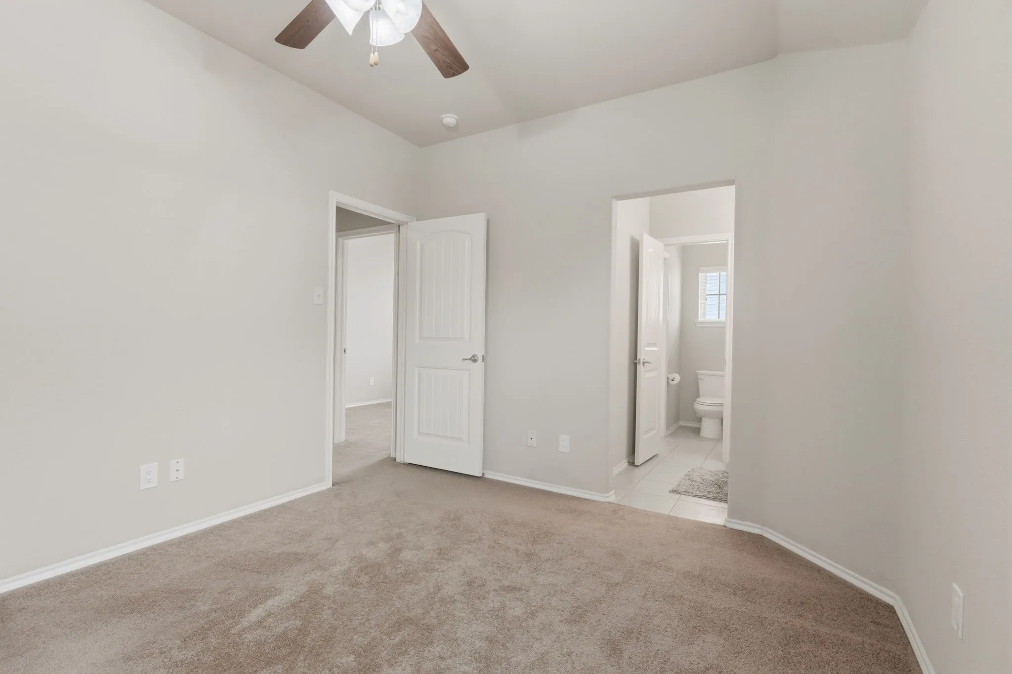 Neutral carpet flooring, white painted walls, ceiling fan with light fixture, white interior doors, and a tiled bathroom floor