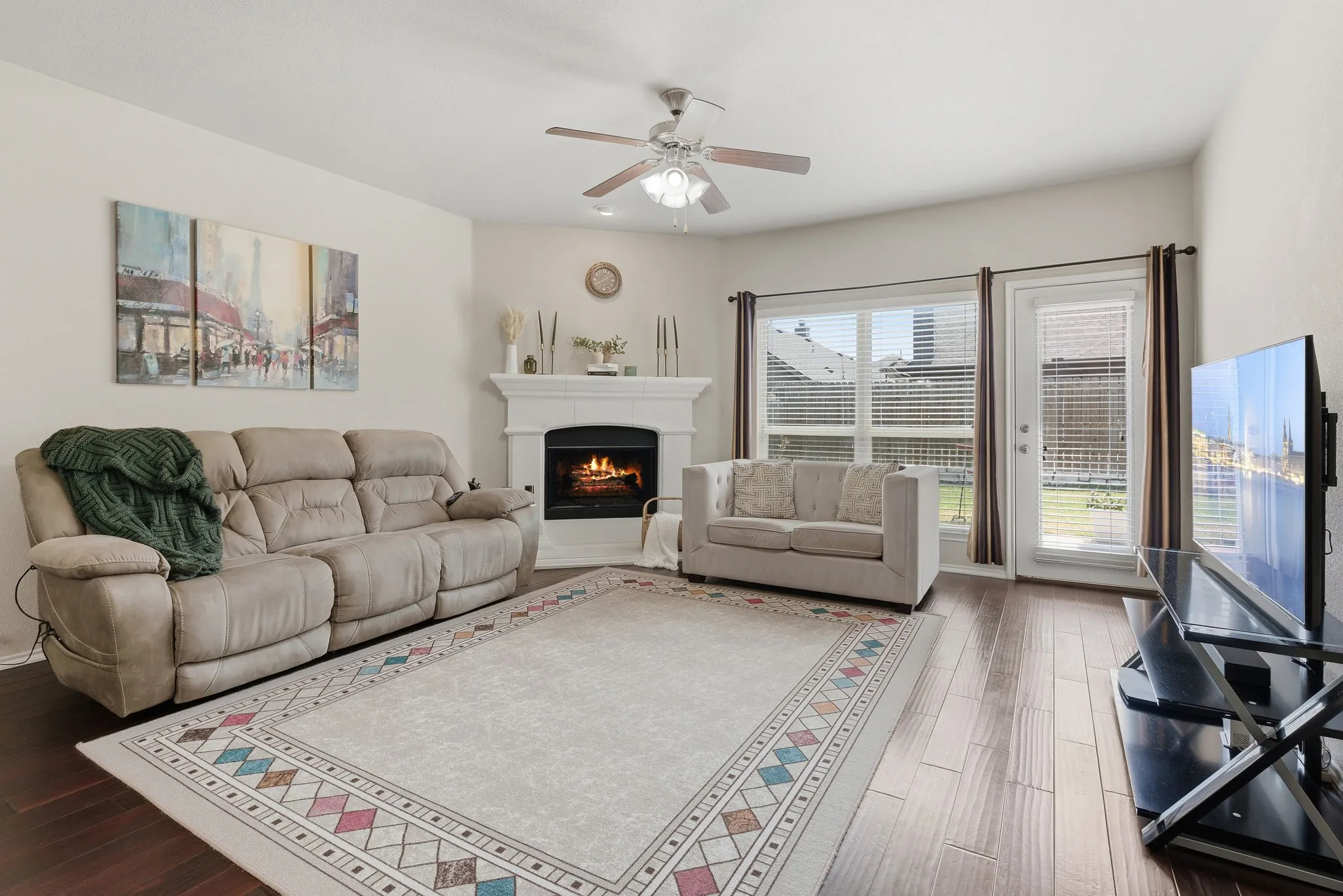 Wood-finish flooring, white mantel fireplace, ceiling fan with integrated lighting, glass-paned door, and large window with blinds