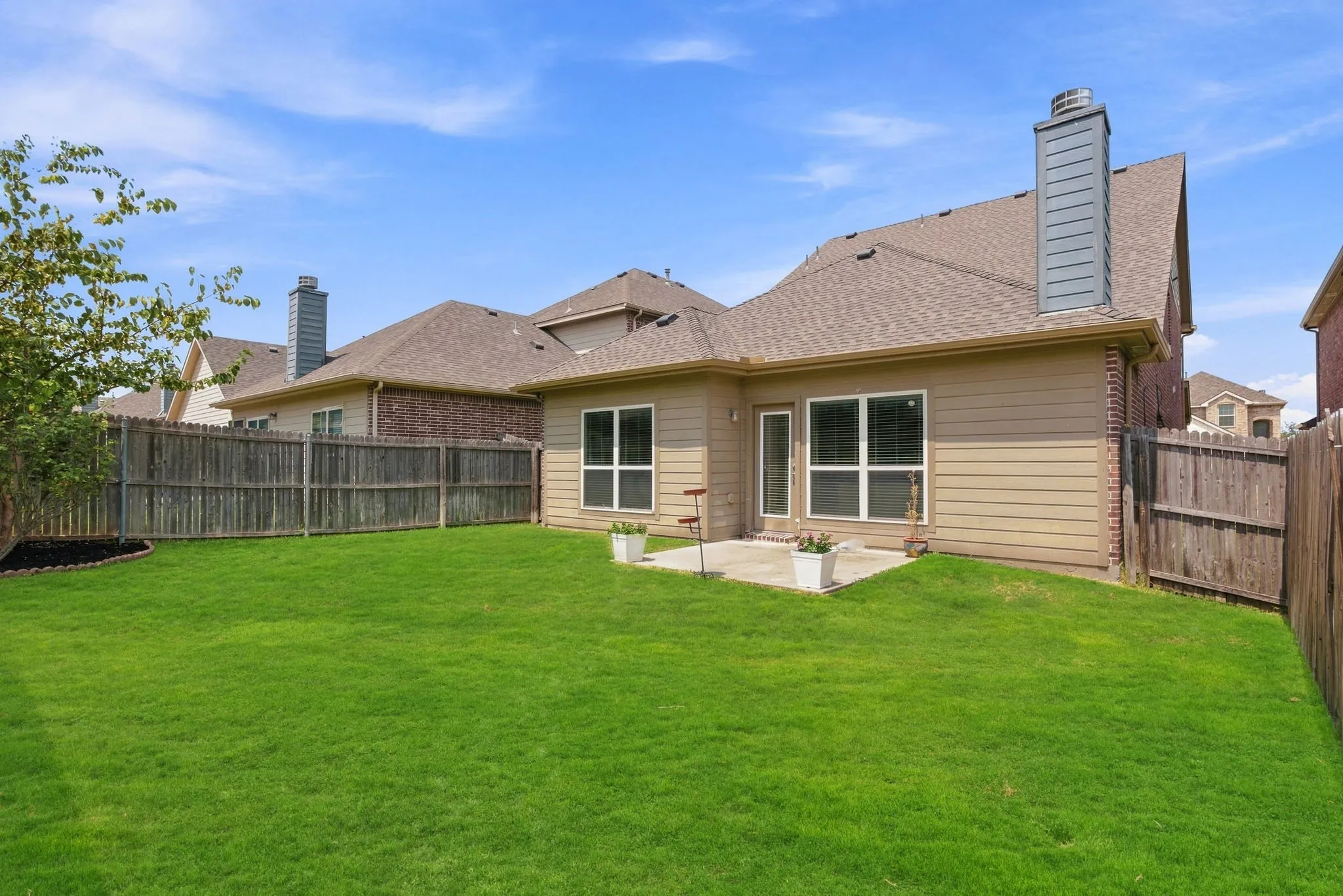 Expansive green lawn, concrete patio, tan siding, brick accents, and a privacy fence