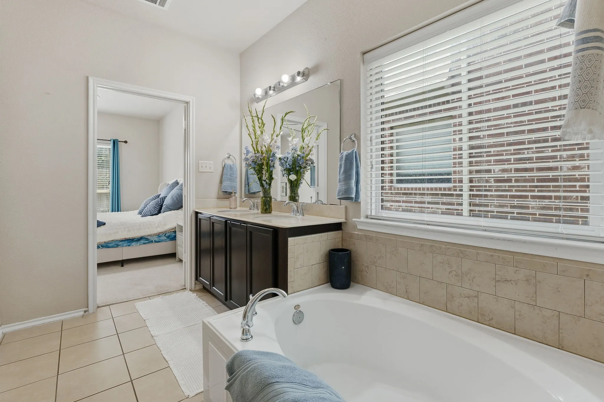 Ceramic tile flooring, white soaking tub, dark wood-finish dual vanity with undermount sinks, and a large window with blinds