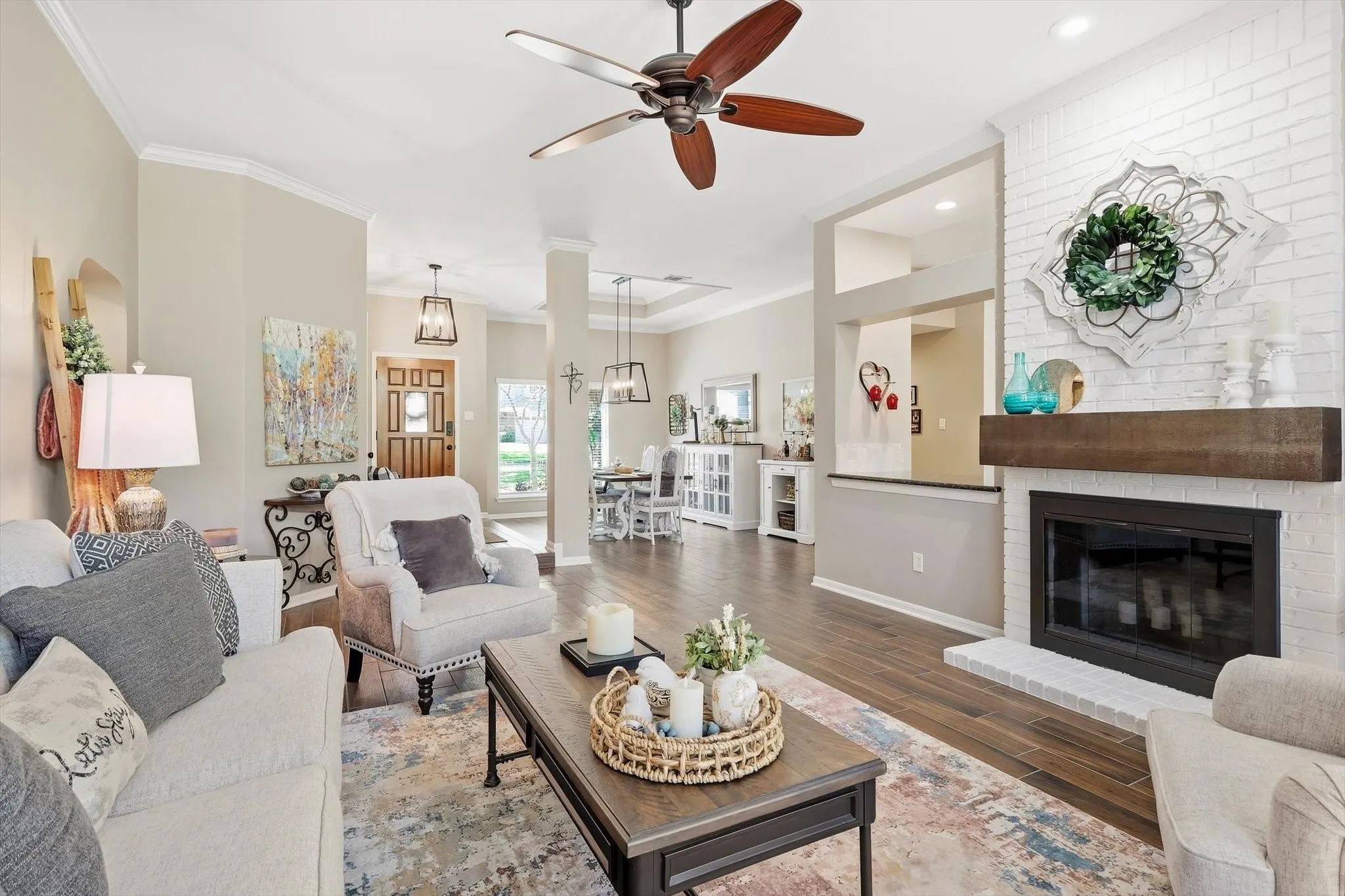 Wood-finish flooring, white brick fireplace with a wood mantel, crown molding, a ceiling fan with light, and a wooden front door