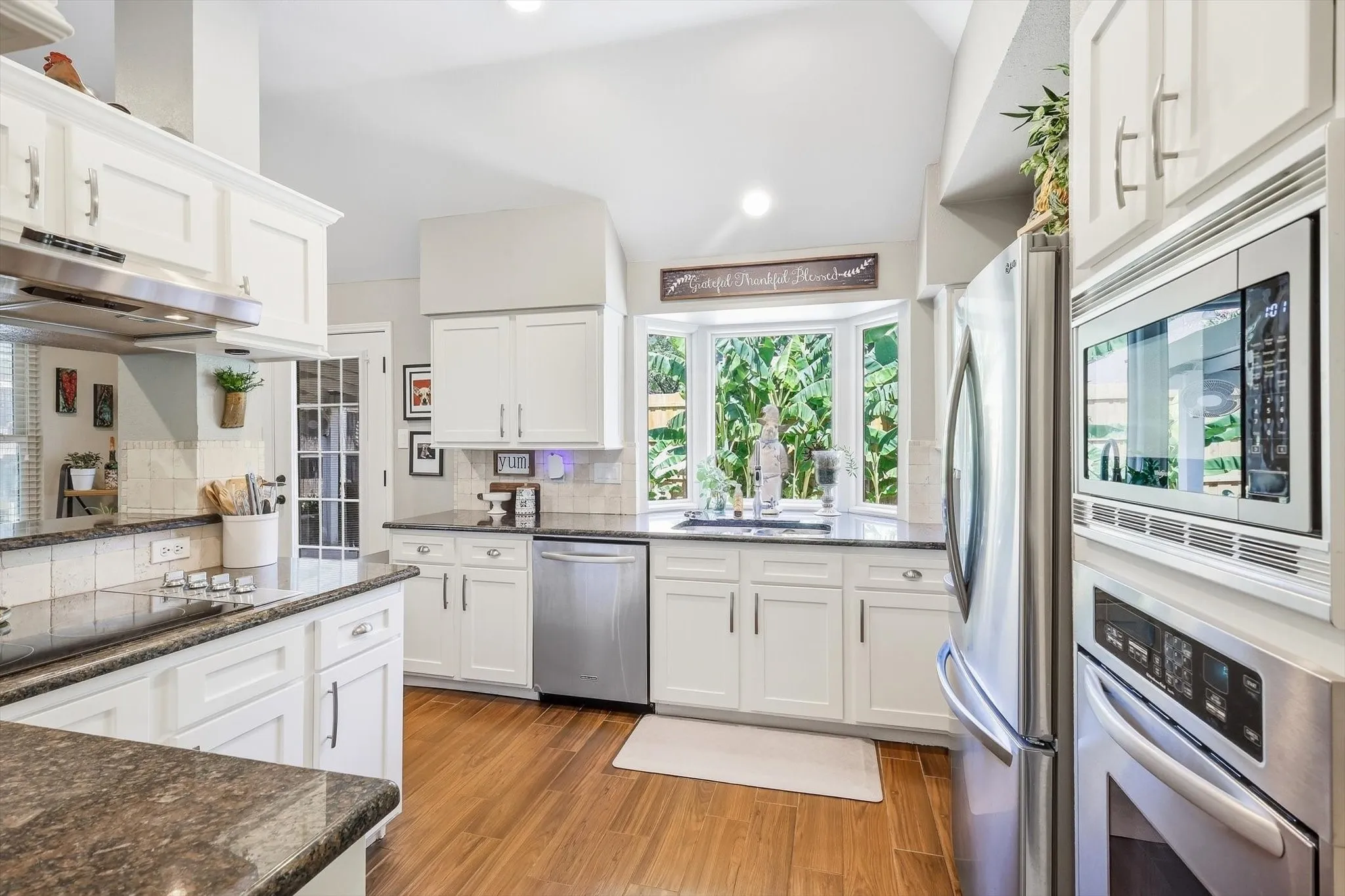 White cabinetry, stainless steel appliances, wood-finish flooring, vaulted ceiling, and a bay window