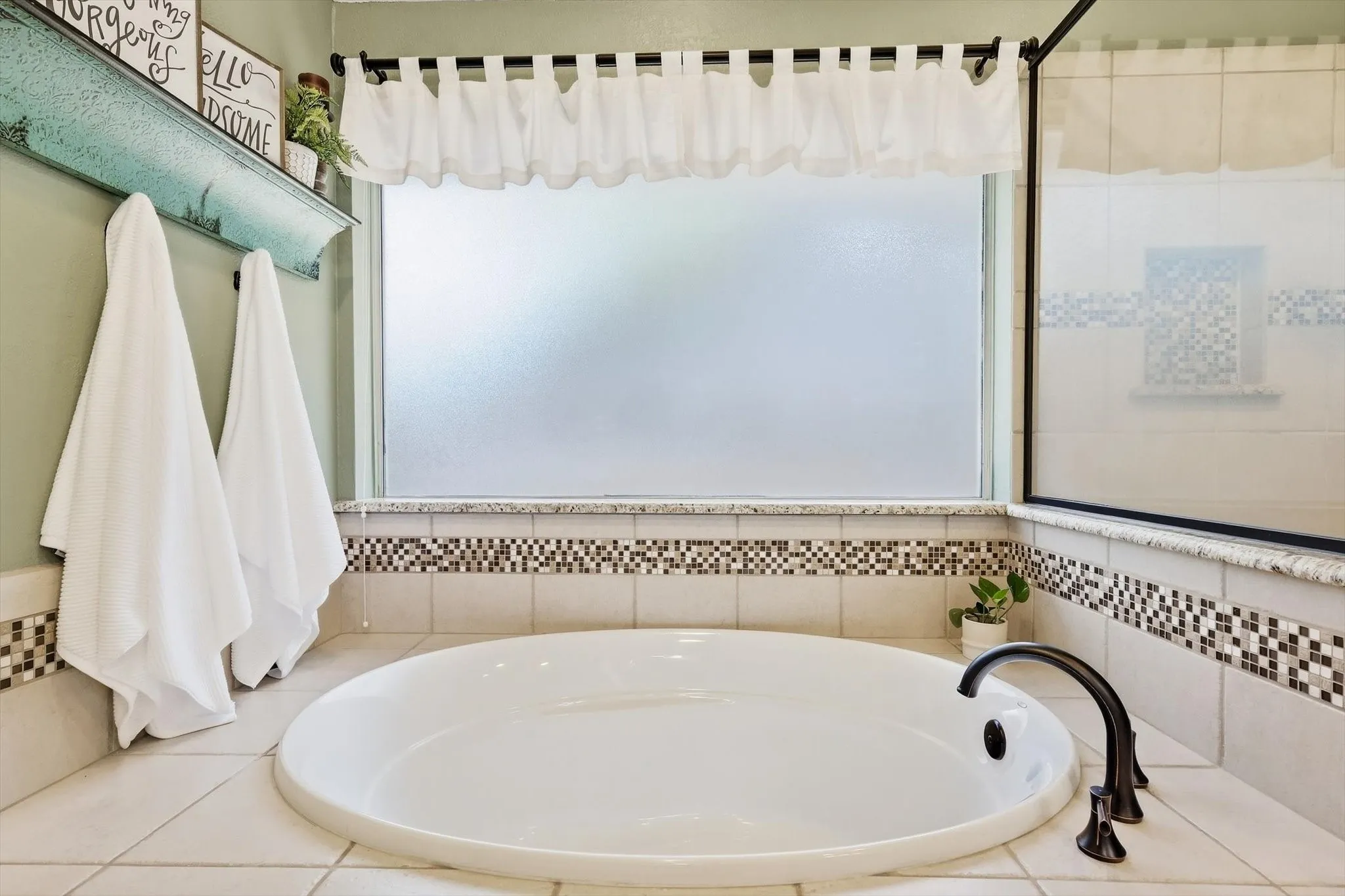 Soaking tub with black-finish fixtures, surrounded by tile and mosaic accents, under a large frosted window