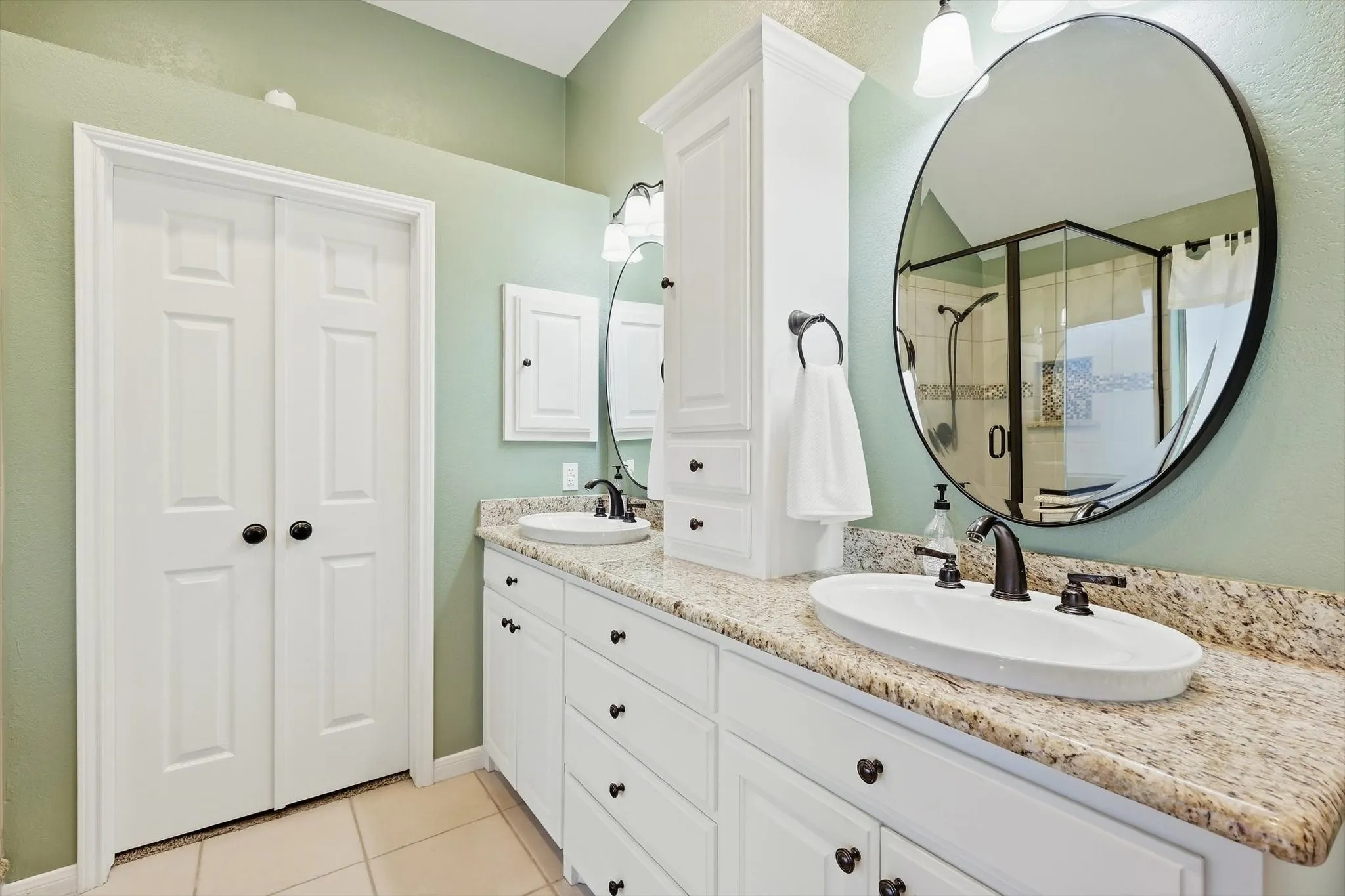 White shaker cabinetry, dual sinks with black fixtures, stone countertop, round mirrors, and a glass-enclosed shower