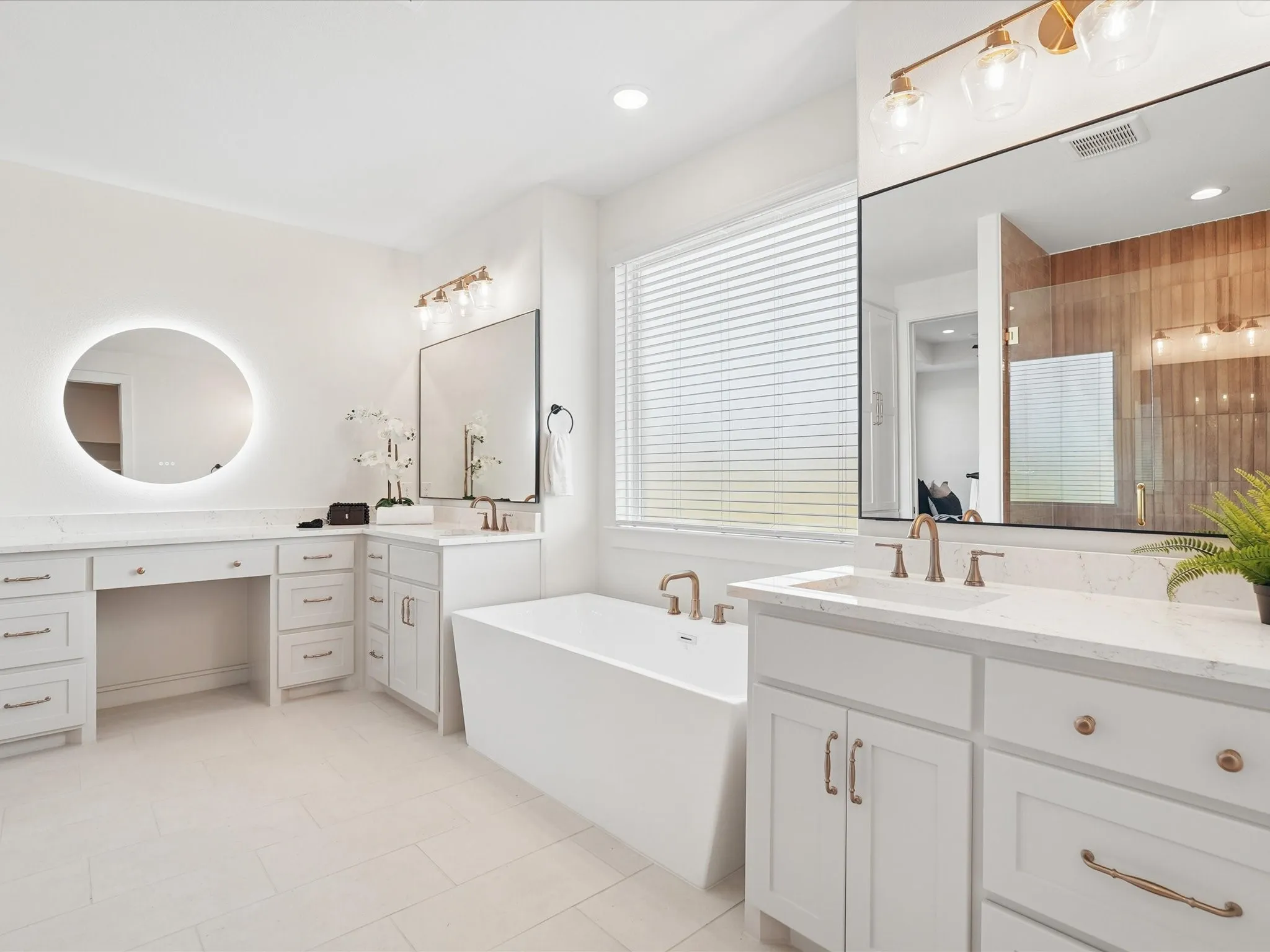 Freestanding soaking tub with floor-mounted faucet, white cabinetry with gold-tone hardware, dual vanity with undermount sinks, and a backlit circular mirror