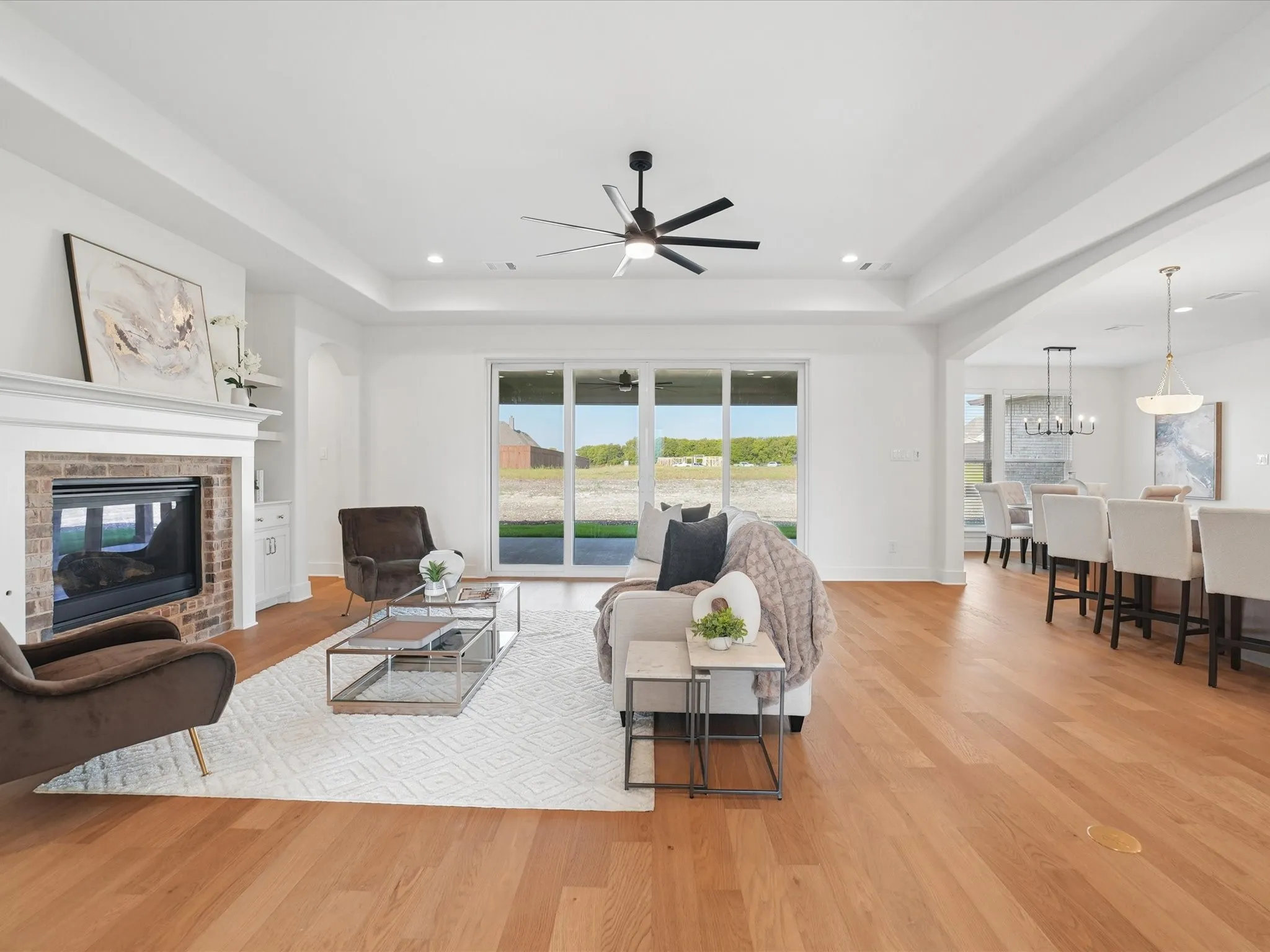 Spacious living area featuring light wood-finish flooring, a brick fireplace with a white mantel, and a tray ceiling with a ceiling fan
