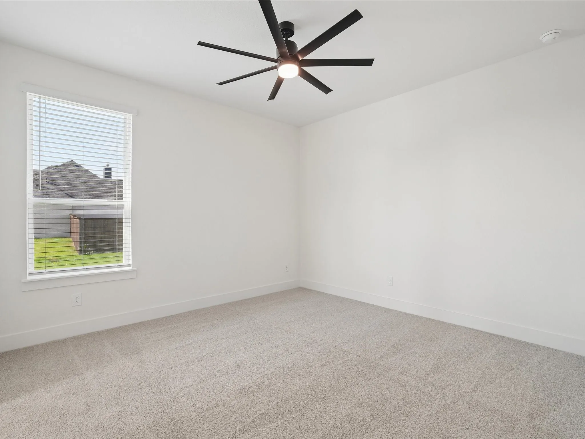 Spacious room featuring neutral carpeting, a large window with blinds, and a modern ceiling fan with integrated lighting