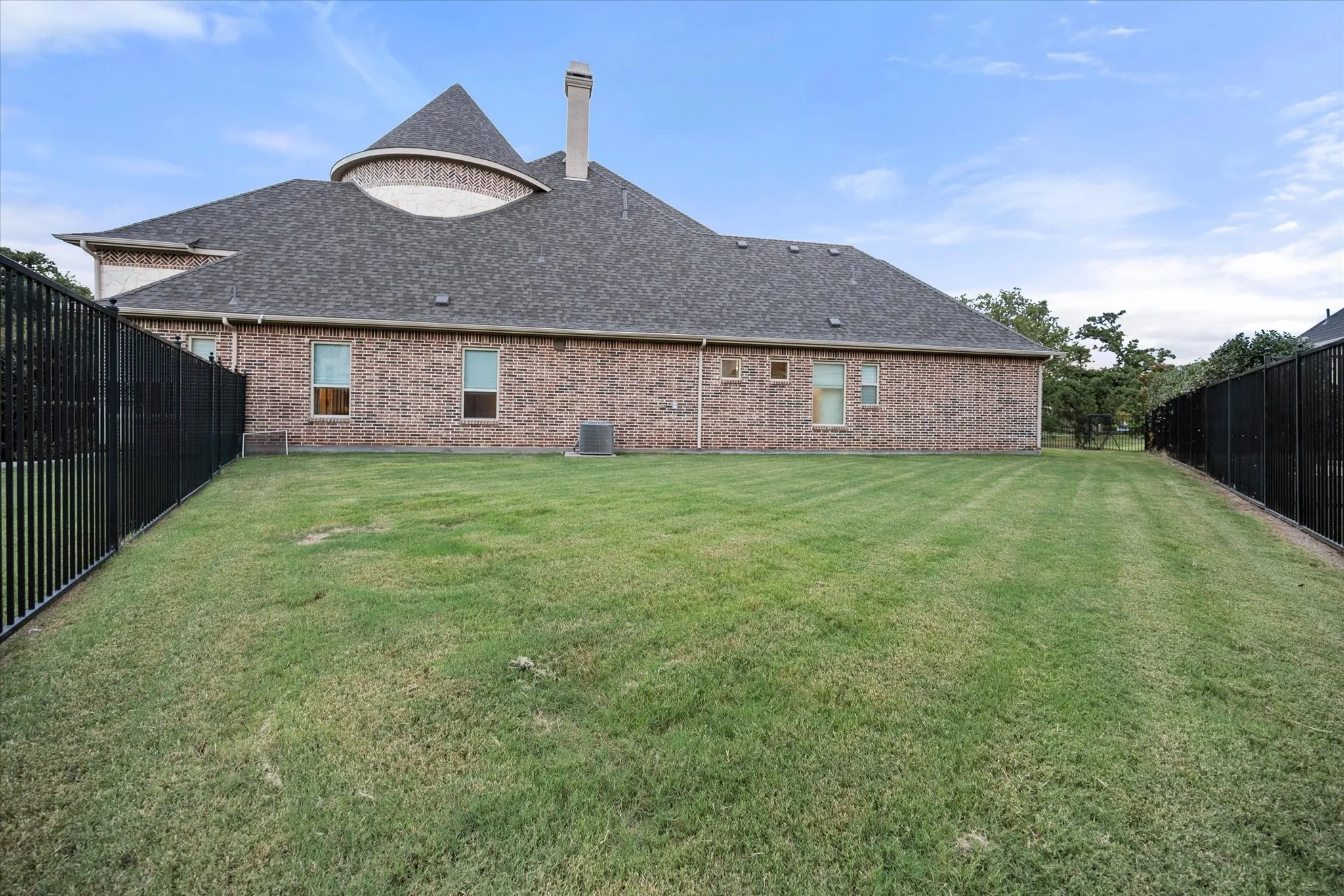 Expansive grass lawn bordered by a black metal fence, featuring a brick exterior with multiple windows and a shingled roof
