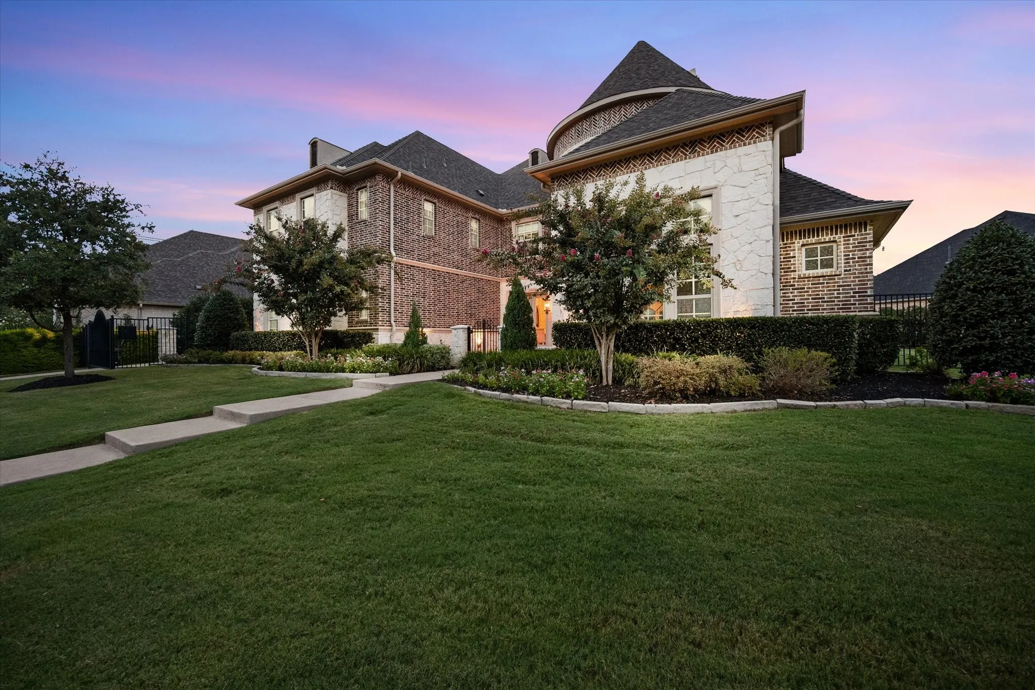 Exterior showcasing brick and stone facade, multi-level roofline, manicured landscaping, concrete pathways, and an iron fence
