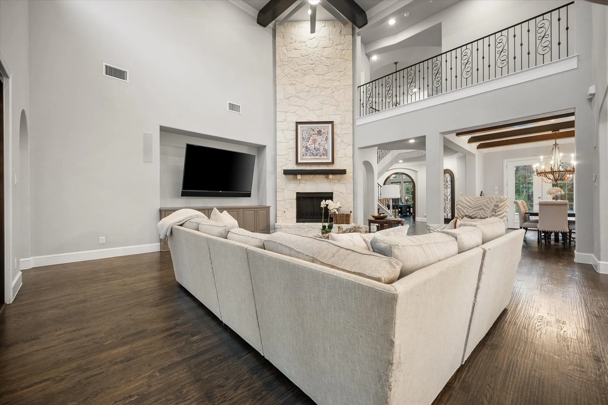 Vaulted ceiling with exposed wood beams, stone fireplace with mantel, dark wood-finish flooring, built-in media cabinet, and iron baluster balcony
