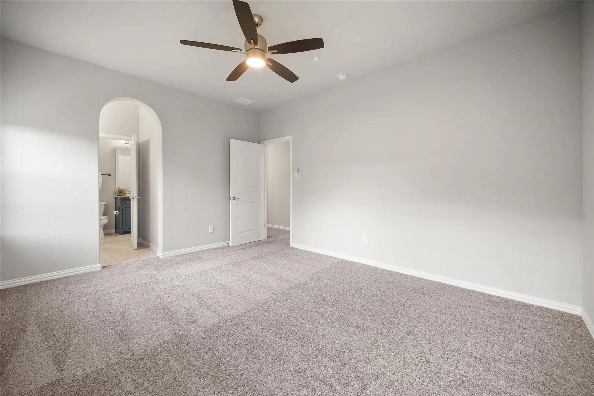 Spacious room featuring neutral carpet flooring, light gray walls, a ceiling fan with integrated lighting, and an arched doorway