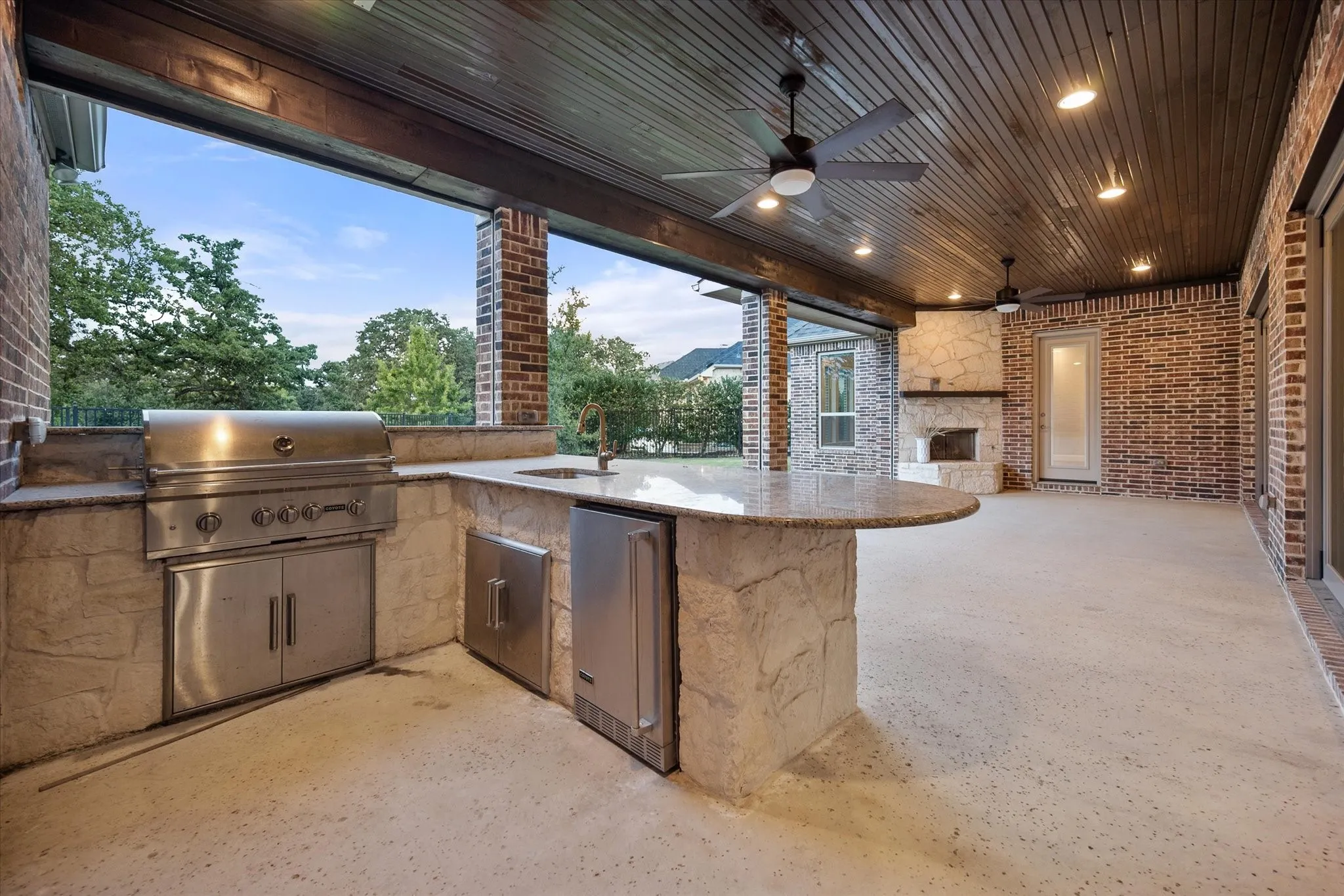 Covered patio featuring an outdoor kitchen with built-in stainless steel grill and sink, a stone-clad fireplace, wood-plank ceiling, and recessed lighting