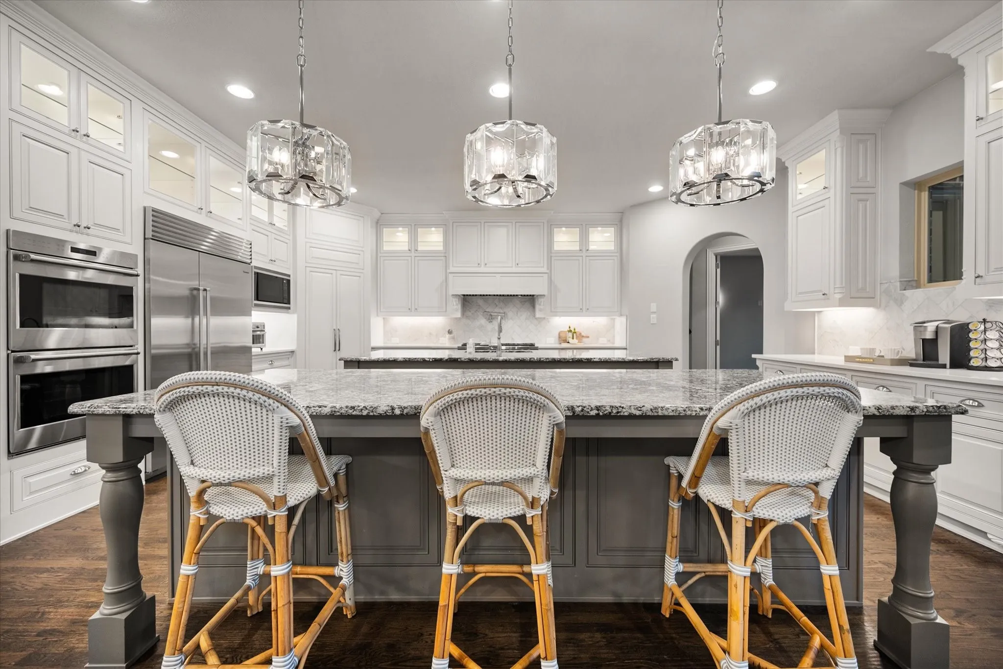 Expansive kitchen featuring a large island with a stone countertop, white cabinetry with glass upper doors, stainless steel double ovens, and a built-in refrigerator