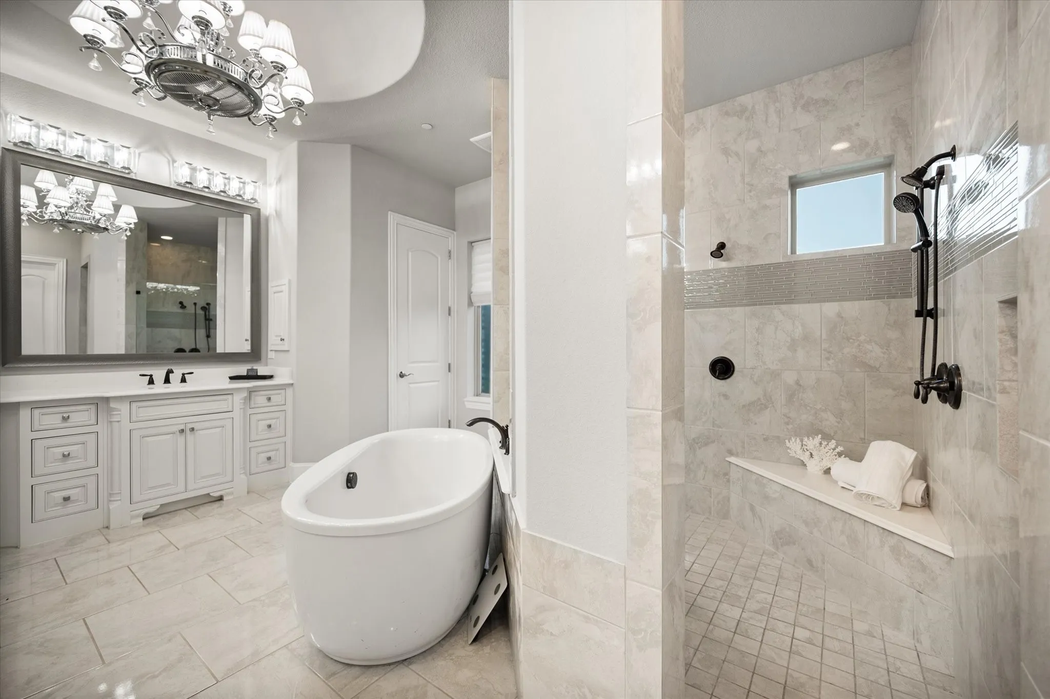 Freestanding soaking tub, walk-in shower with bench, dual vanity with extensive storage, recessed shower niche, and ceiling chandelier