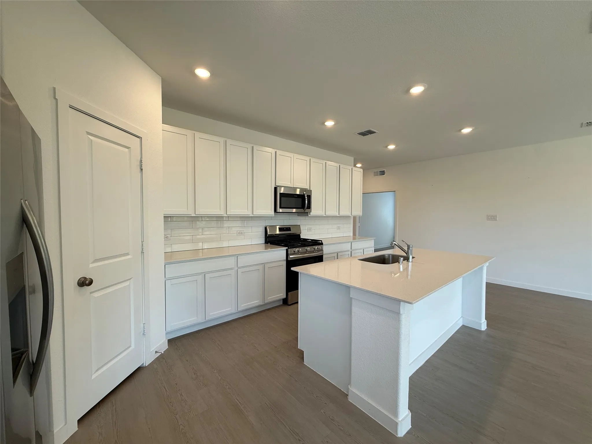 Open-concept kitchen featuring white cabinetry, stainless steel appliances, a center island with an integrated sink, recessed lighting, and wood-finish flooring