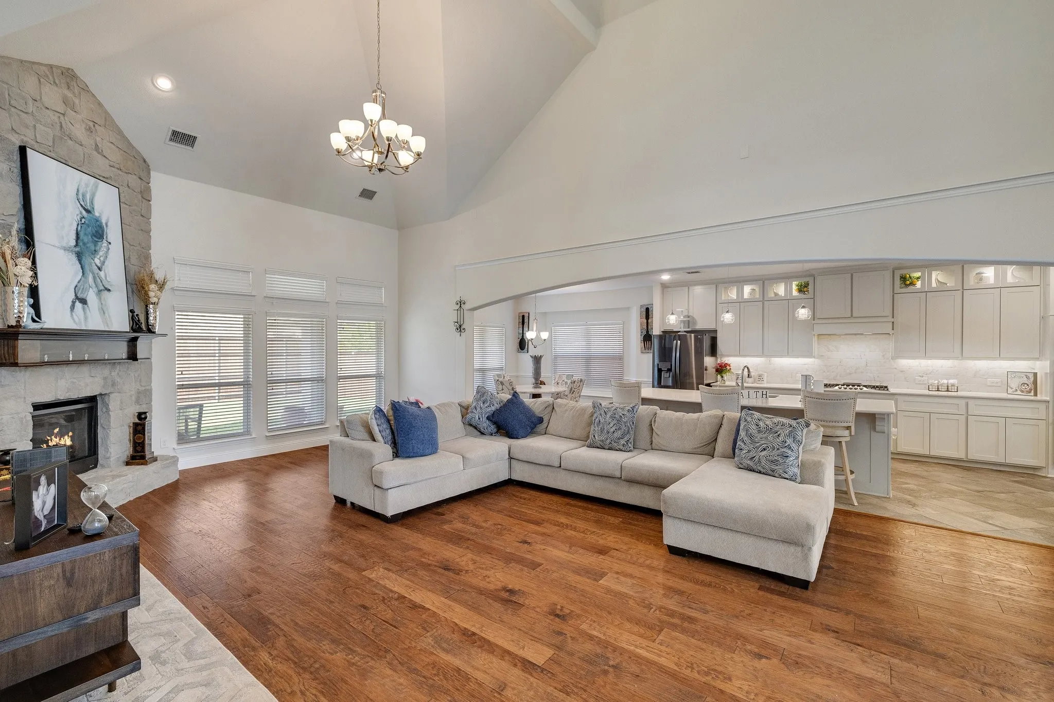 Vaulted ceiling with chandelier, stone-clad fireplace, wood-finish flooring, multiple windows, and a kitchen with white cabinetry and island seating