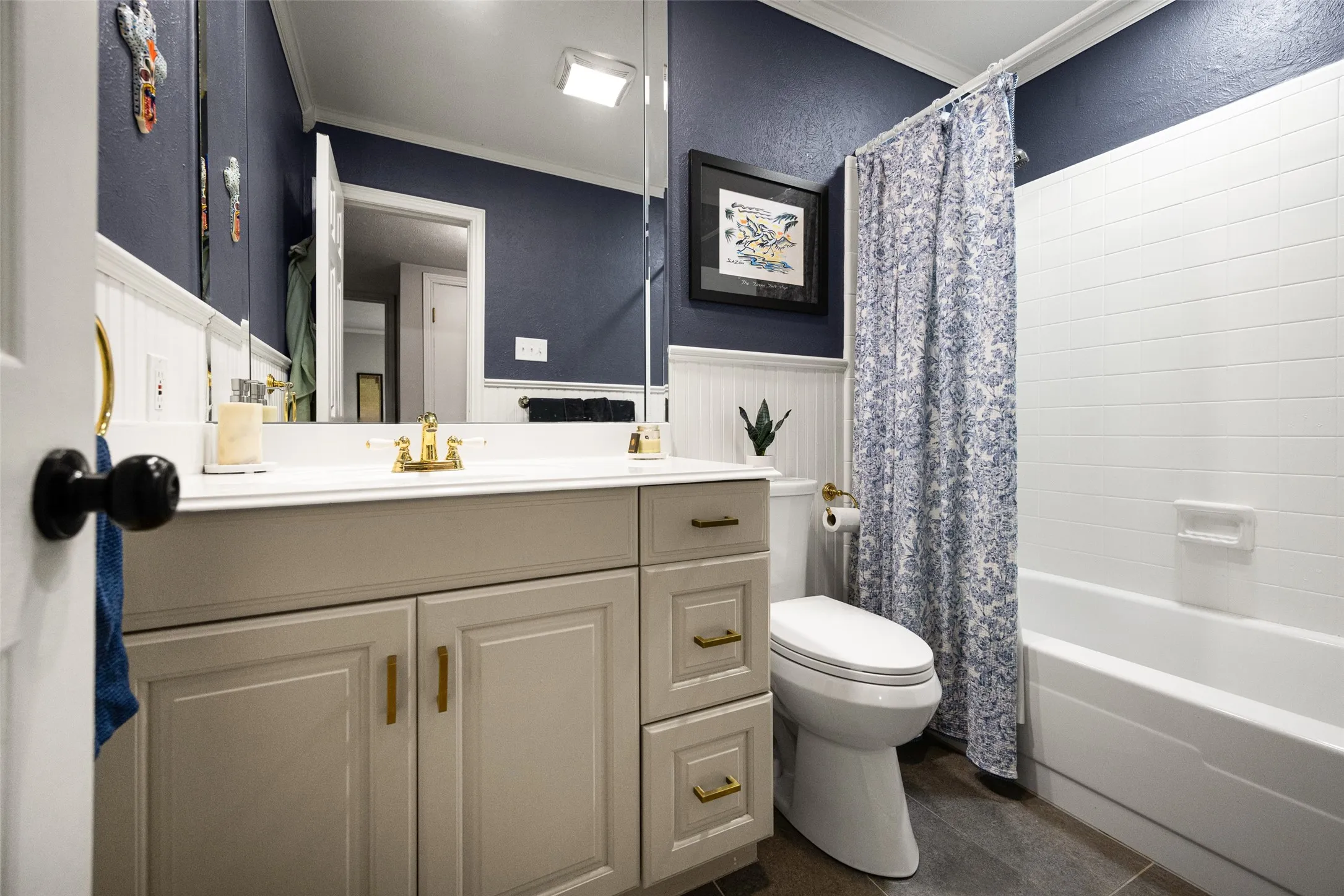 Vanity with gold-tone fixtures, wood-look flooring, white paneled wall detailing, and a white tiled shower surround