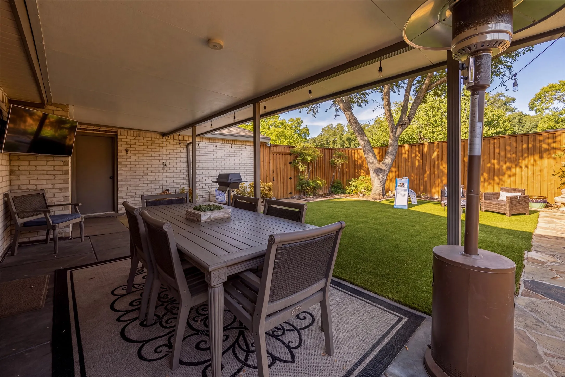 Covered patio featuring string lighting, brick exterior wall, and an expansive lawn with mature trees and wood fencing