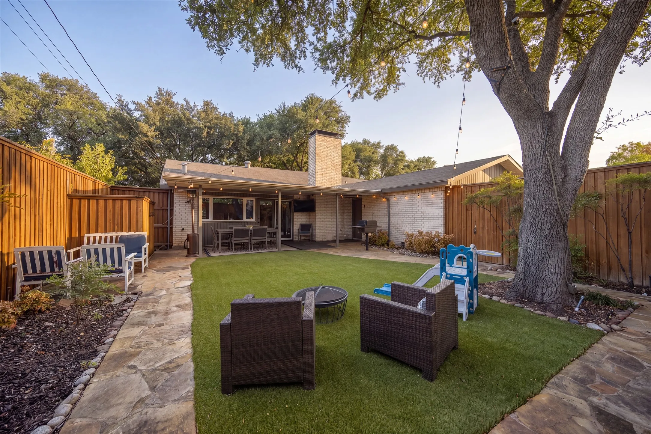 Manicured backyard featuring a stone patio, artificial turf, a mature tree, and privacy fencing