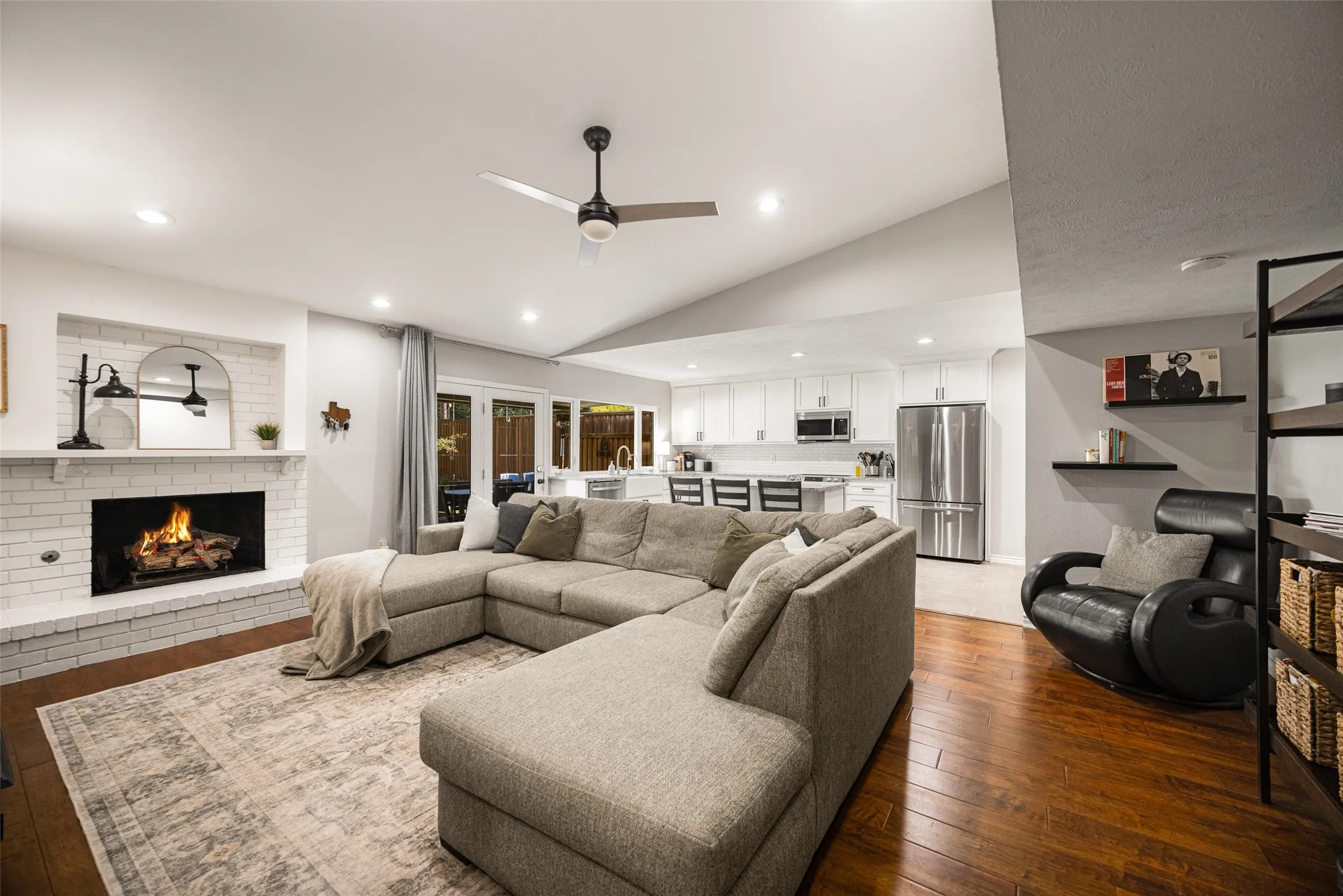 Open-concept living space featuring wood-finish flooring, a white brick fireplace, vaulted ceilings, recessed lighting, and a modern ceiling fan