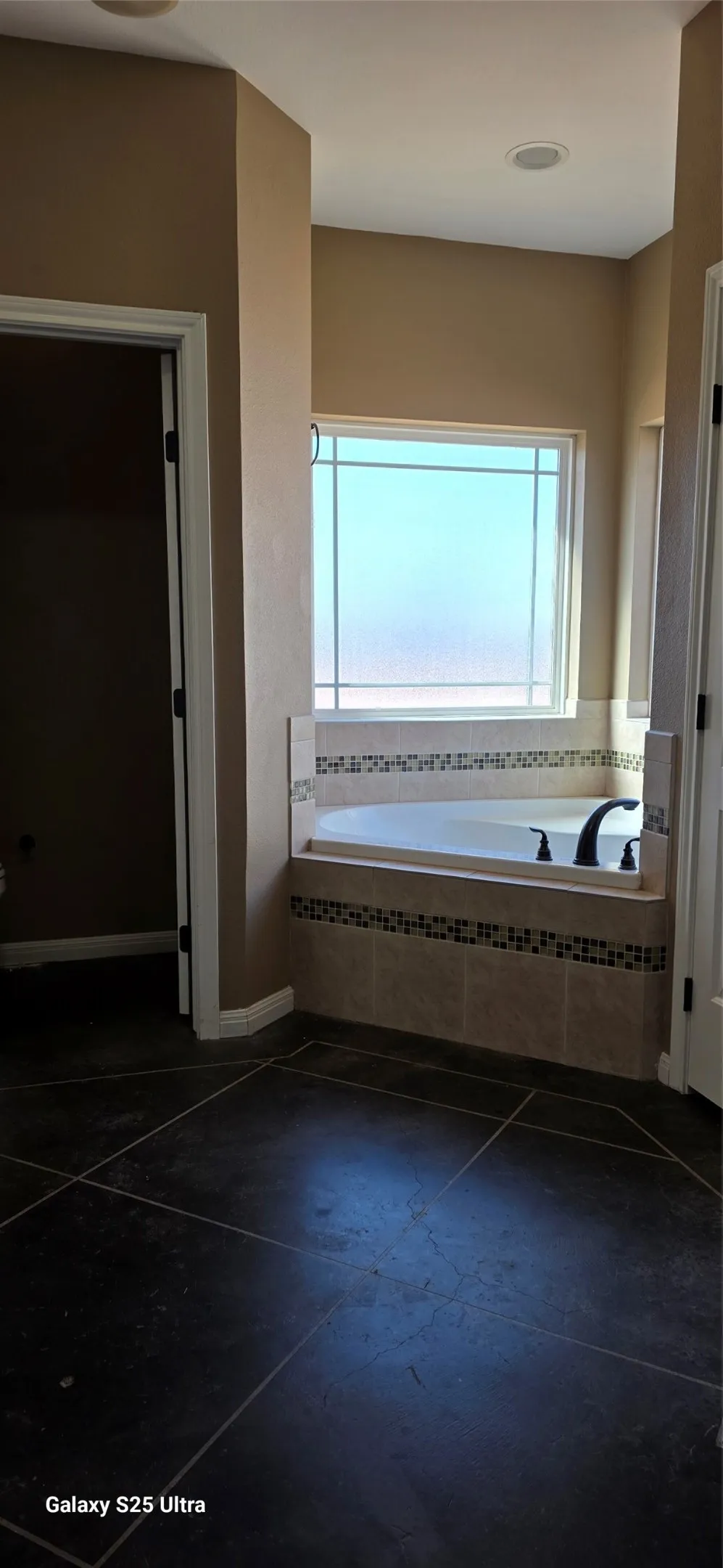 Soaking tub with mosaic tile surround, frosted window, recessed lighting, and dark tile flooring