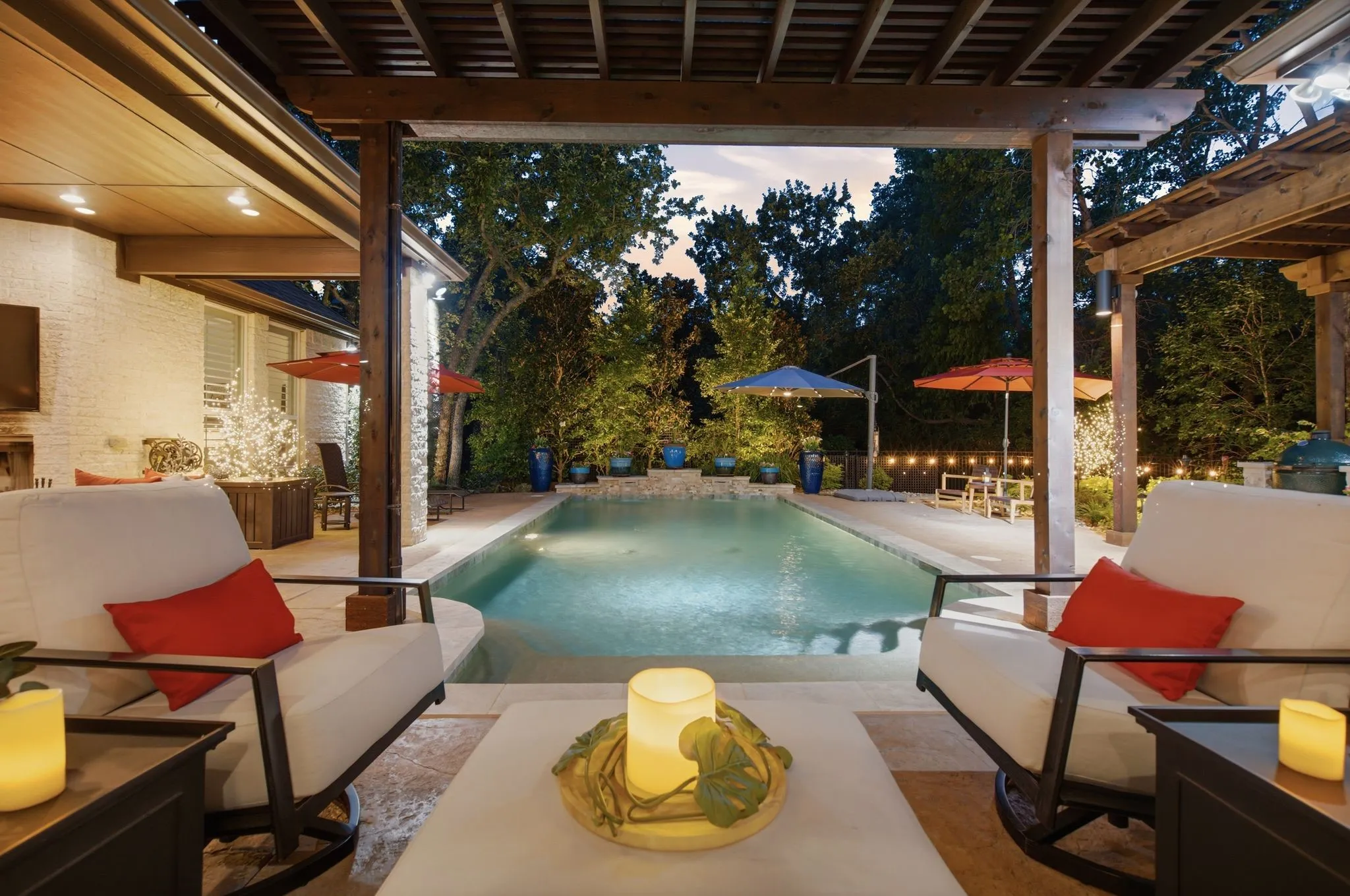Step outside and settle in—this inviting view from the covered outdoor living area captures the backyard at its best. The sparkling pool stretches toward a beautifully landscaped backdrop of mature trees, while multiple poolside seating areas and warm evening lighting create an intimate, resort-like setting. Designed for both entertaining and unwinding, this is outdoor living meant to be enjoyed from morning coffee through evenings under the stars.