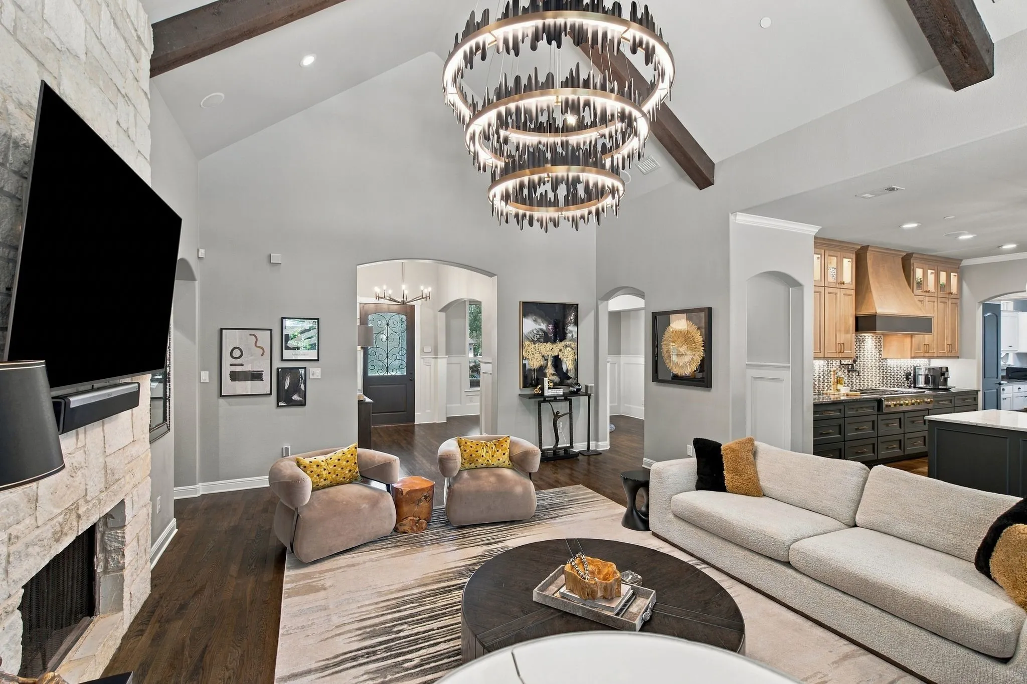 This photo shows the scale of the great room and its connection to the kitchen and foyer. A sweeping view of the great room showcases the dramatic vaulted ceiling, exposed wood beams, statement chandelier and floor-to-ceiling stone fireplace. Graceful arched openings, rich hardwood floors and an open connection to the beautifully appointed kitchen create a sophisticated yet comfortable space designed for effortless entertaining.