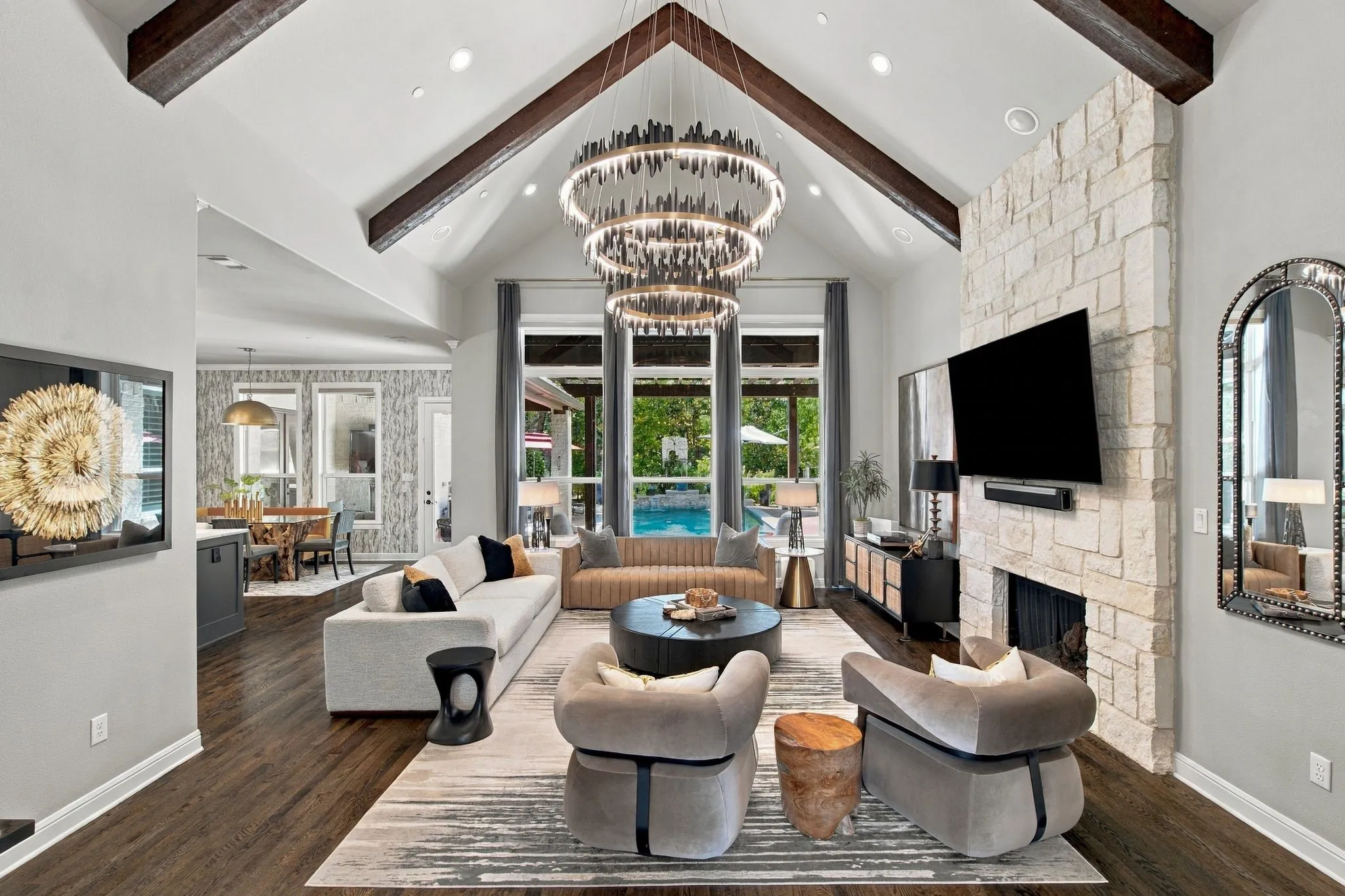 Dramatic great room featuring a soaring vaulted ceiling with rich exposed beams, a floor-to-ceiling stone fireplace and striking designer lighting. Expansive windows fill the room with natural light and frame beautiful views of the pool and outdoor living area. The sophisticated interiors, including art, furnishings, décor and lighting, were thoughtfully curated by Sally Lynn Home of Southlake.