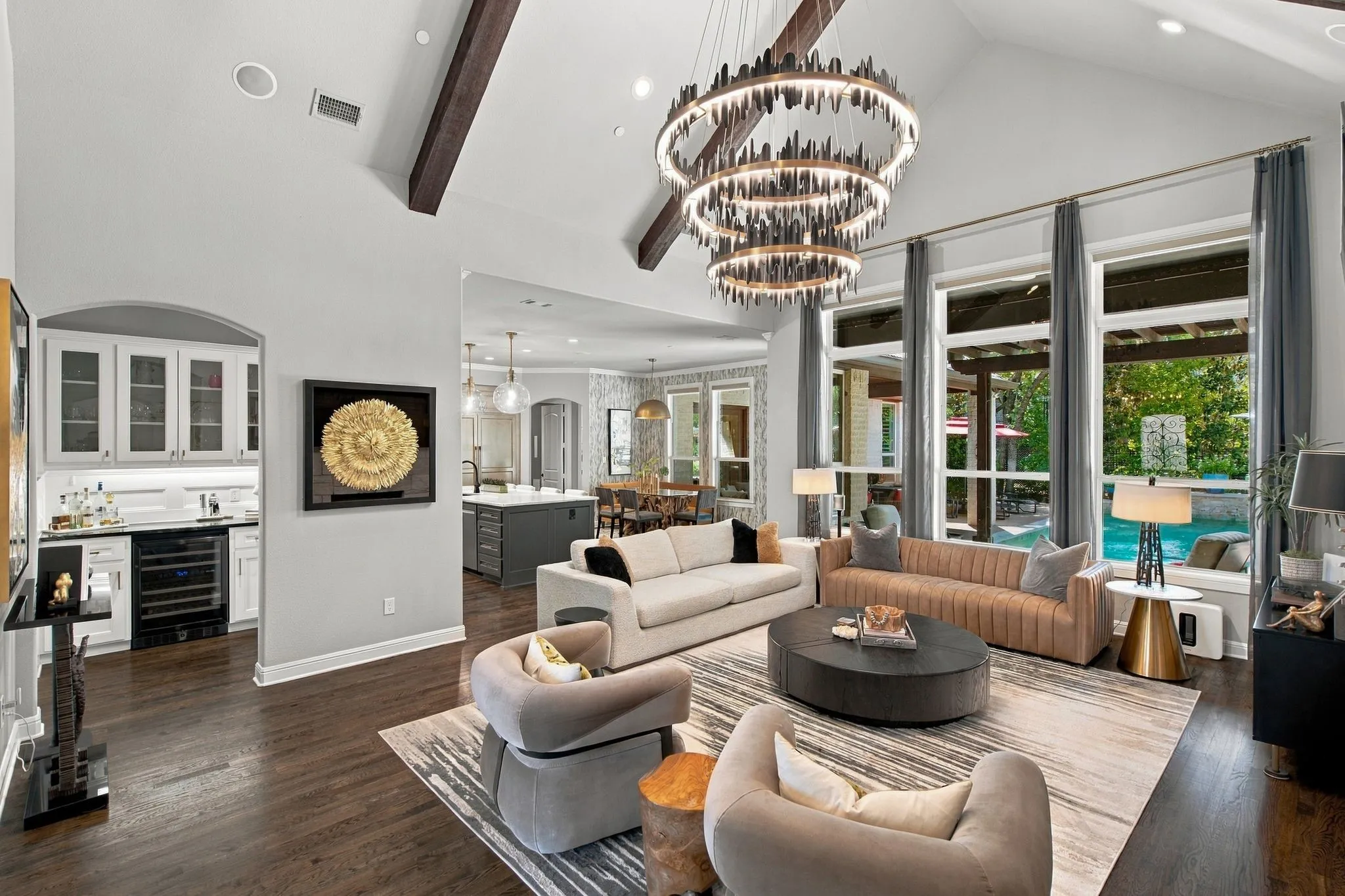 Another view of the impressive great room highlights the soaring beamed ceiling, dramatic designer lighting and rich hardwood floors. Expansive windows overlook the pool and outdoor living area, while the open layout flows effortlessly into the kitchen, casual dining area and built-in beverage bar—an exceptional setting for everyday living and entertaining.