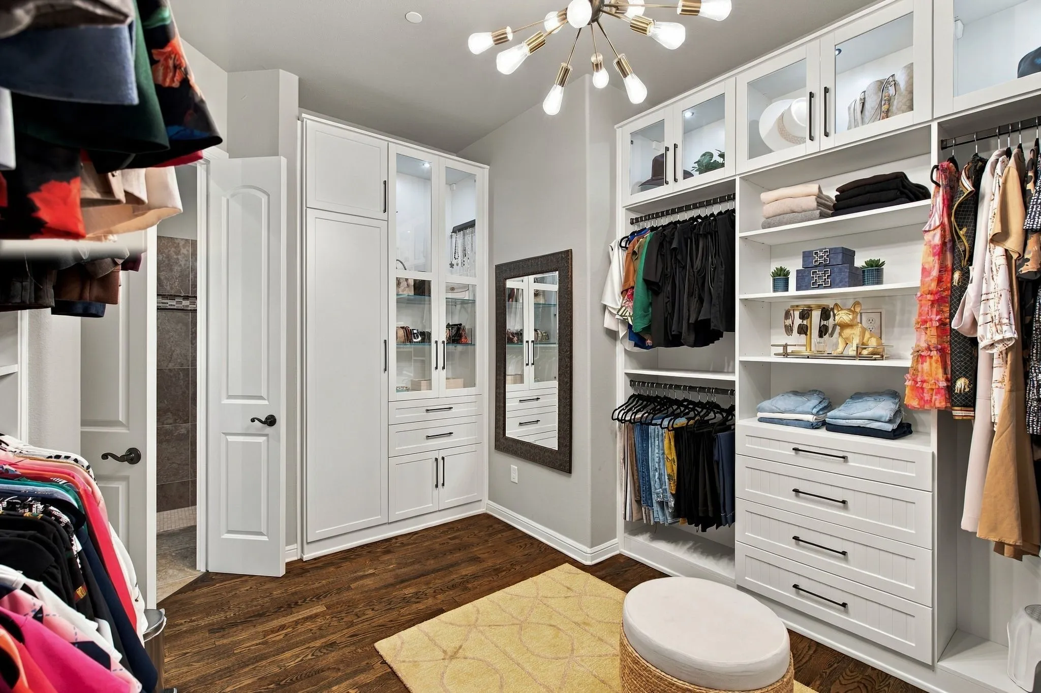 Another view of the custom California primary closet reveals exceptional attention to detail, with floor-to-ceiling built-ins, illuminated glass-front cabinetry, extensive drawer storage and thoughtfully designed hanging and display areas. Beautiful hardwood floors and statement lighting complete this luxurious, boutique-style dressing space.