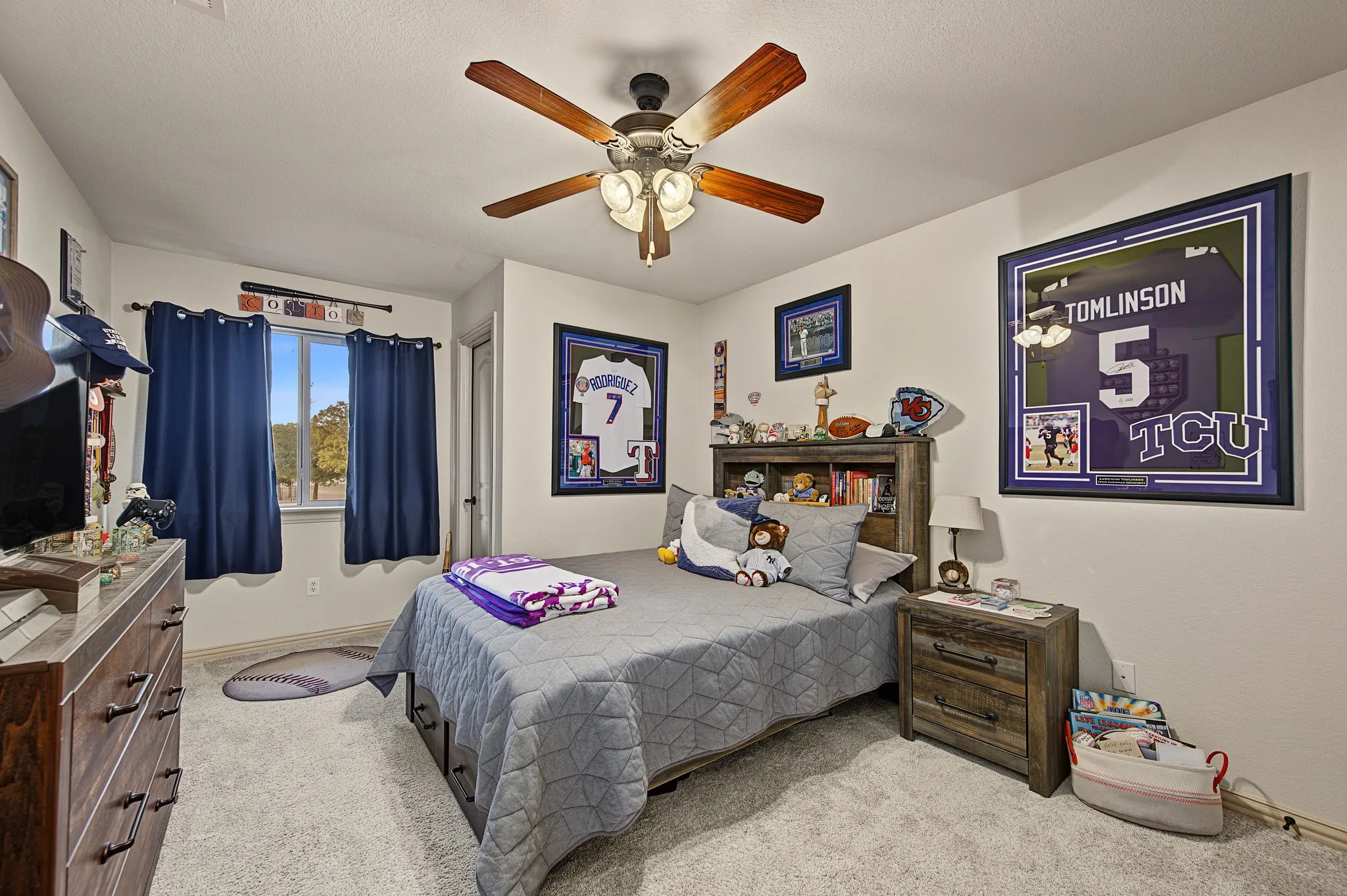 Upstairs bright secondary bedroom featuring a ceiling fan with integrated lighting, carpeted flooring, wood-finish nightstand, and large window with blue curtain treatments