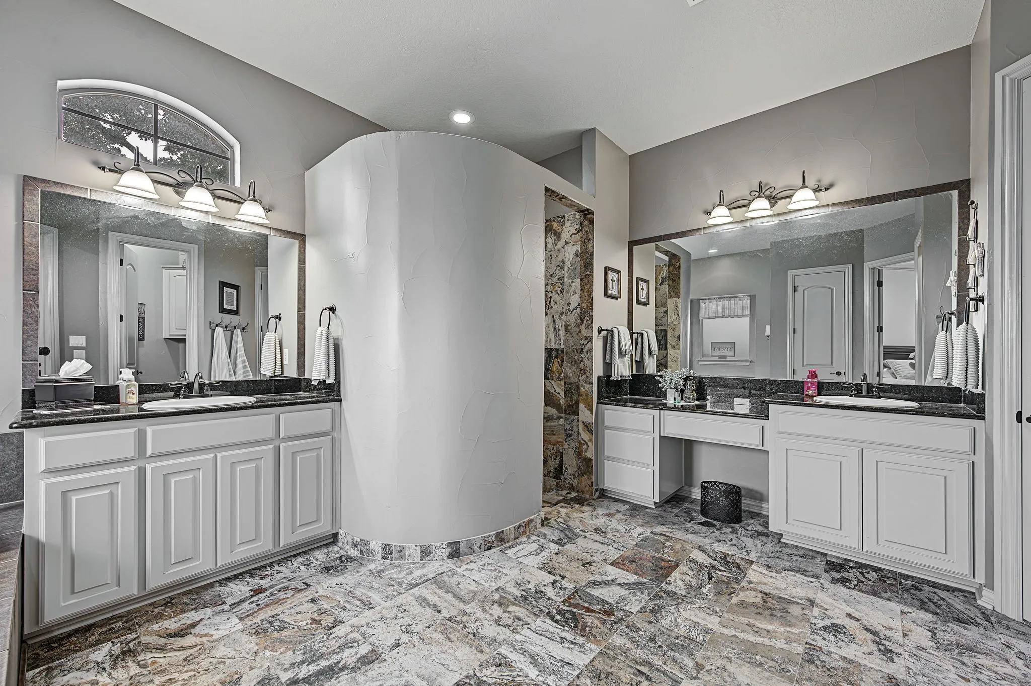 Spacious bathroom with dual vanities, dark granite countertops, built-in makeup area, and large format floor tiling