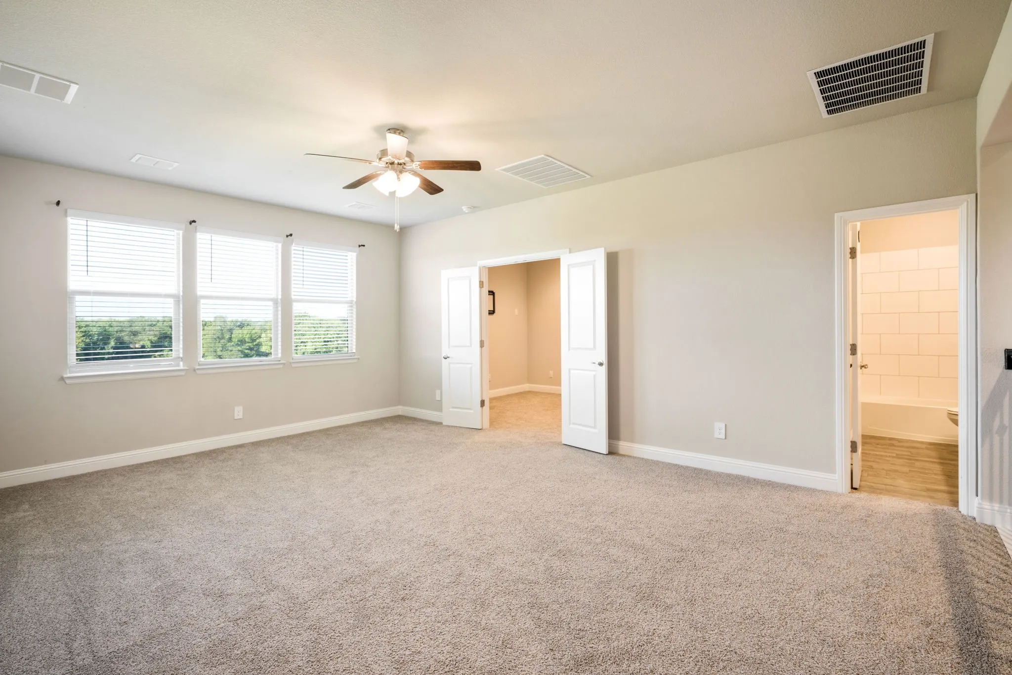 Spacious room with carpeted flooring, multiple windows, ceiling fan, and access to an additional room through double doors