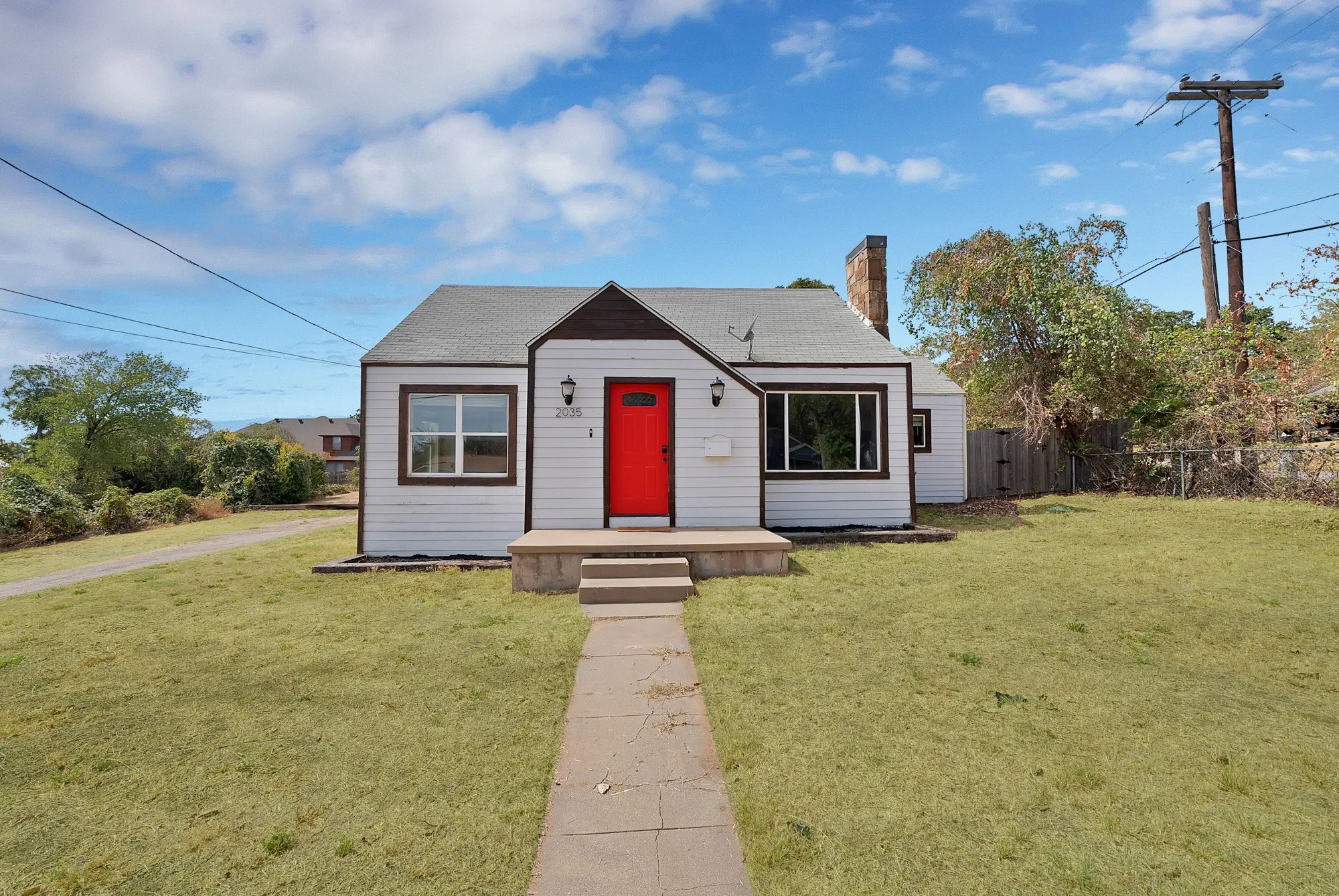 Front exterior: Cape Cod style home with stone chimney, red front door, and dark wood trim accents. Front lawn, concrete walkway, driveway, and wood privacy fence with gate.