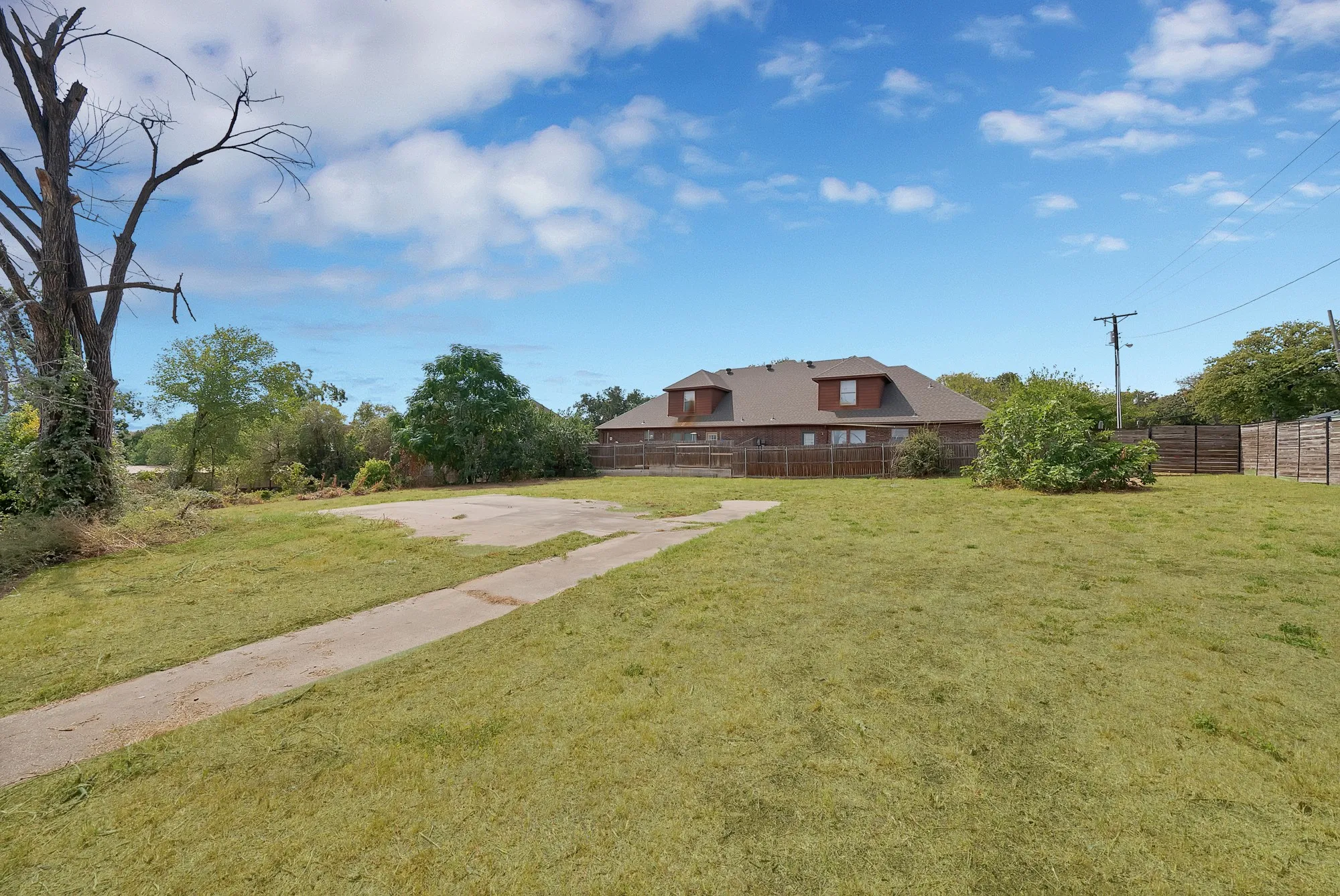 Backyard: Open grass yard with concrete walkway, concrete slab, and wood privacy fencing. Mature trees and shrubs along the property line.