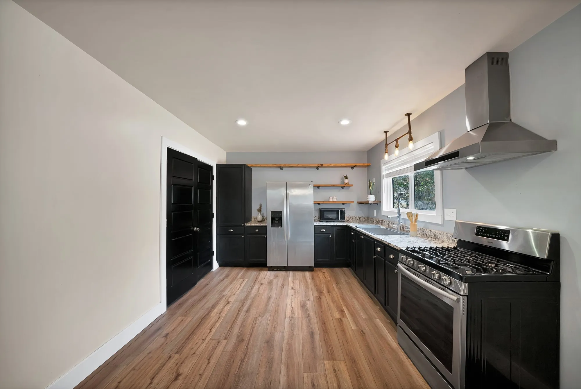 Kitchen: Granite countertops, stainless steel sink, and commercial-style pull-down faucet under a window. Samsung gas range, stainless chimney vent hood, industrial pipe pendant with Edison bulbs, and open wood floating shelves on pipe brackets.