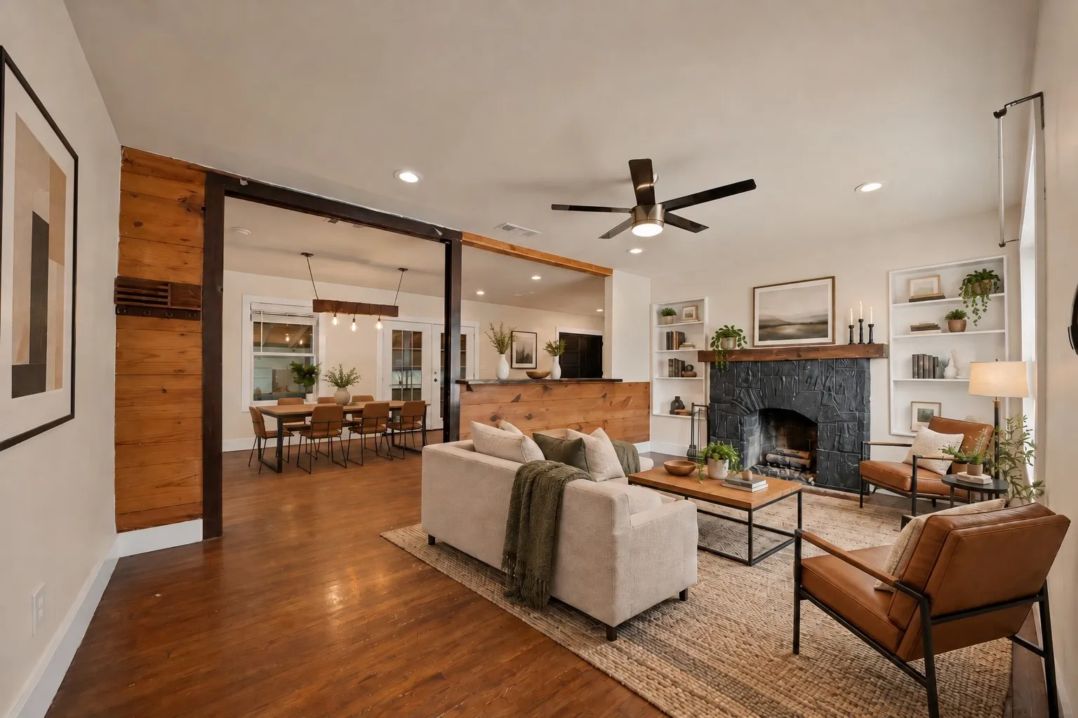virtually staged Living room: Open concept layout with black painted stone fireplace, reclaimed wood beam mantel, and flanking built-in bookshelves. Knotty pine plank breakfast bar, exposed wood beam, ceiling fan, recessed lighting, and hardwood floors open to the dining area with French doors.