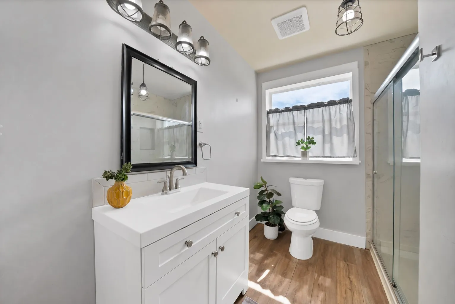 Bathroom: White shaker vanity with integrated sink top, brushed nickel faucet, and subway tile backsplash. Walk-in shower with marble-look tile and sliding glass doors, industrial cage pendant lighting, window, and wood-look plank flooring.